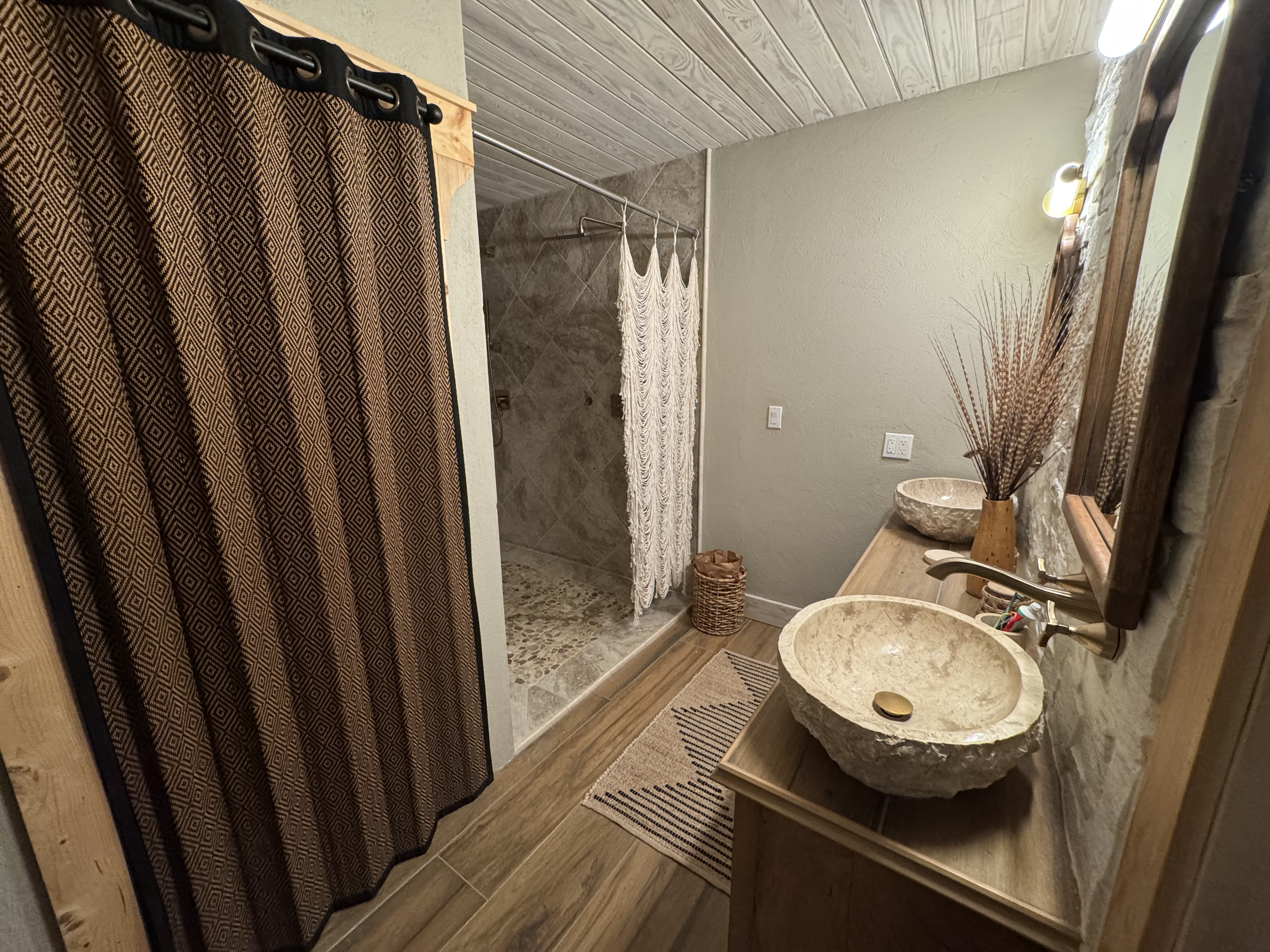 A rustic bathroom with a stone vessel sink on a wooden vanity, a mirror, a vase with dried plants, and a textured wall. The shower area features a pebble floor and a decorative curtain.