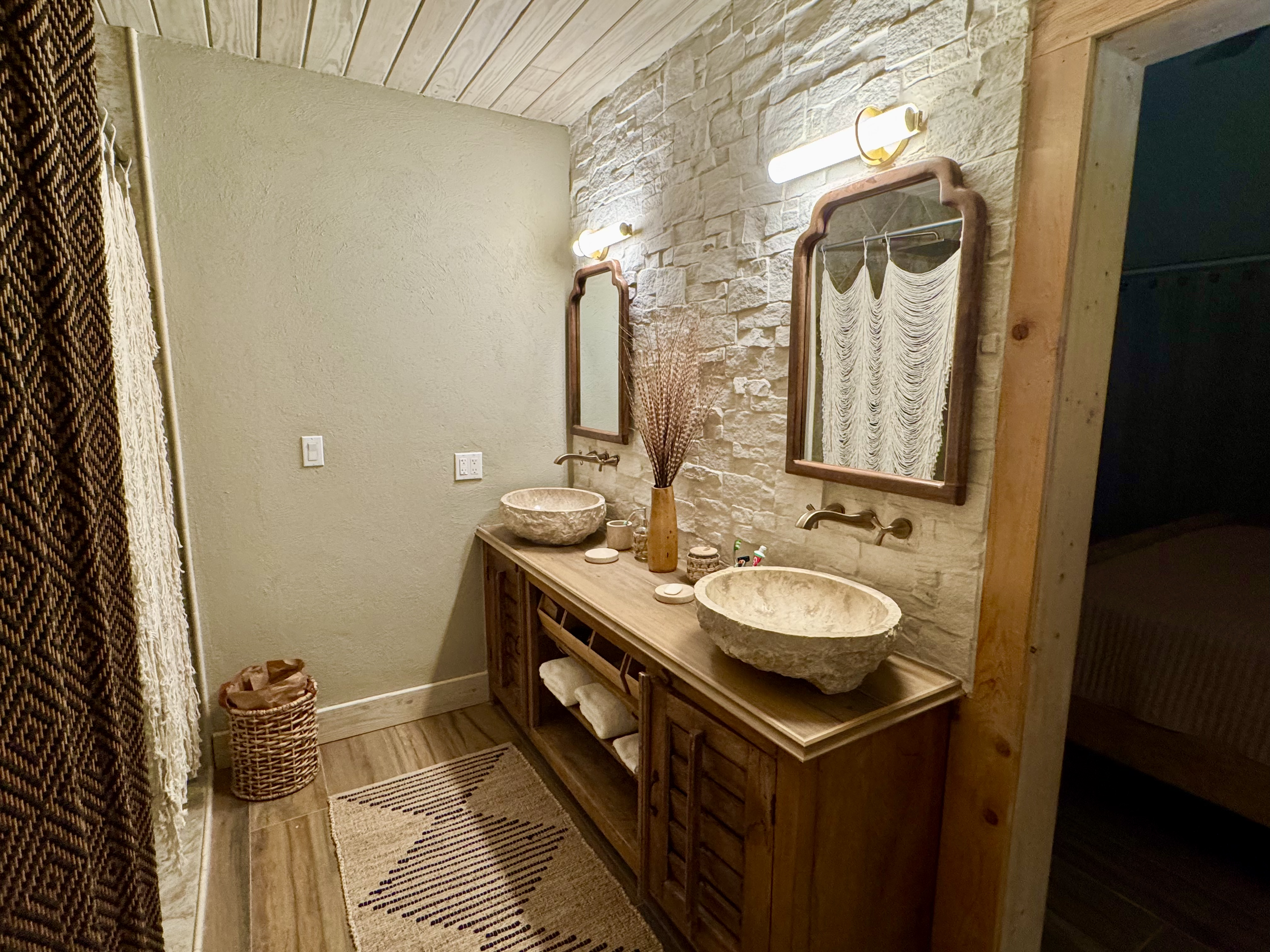 Bathroom with two stone vessel sinks, wooden vanity, mirrors, wall lights, a woven basket, a potted plant, and a striped rug.
