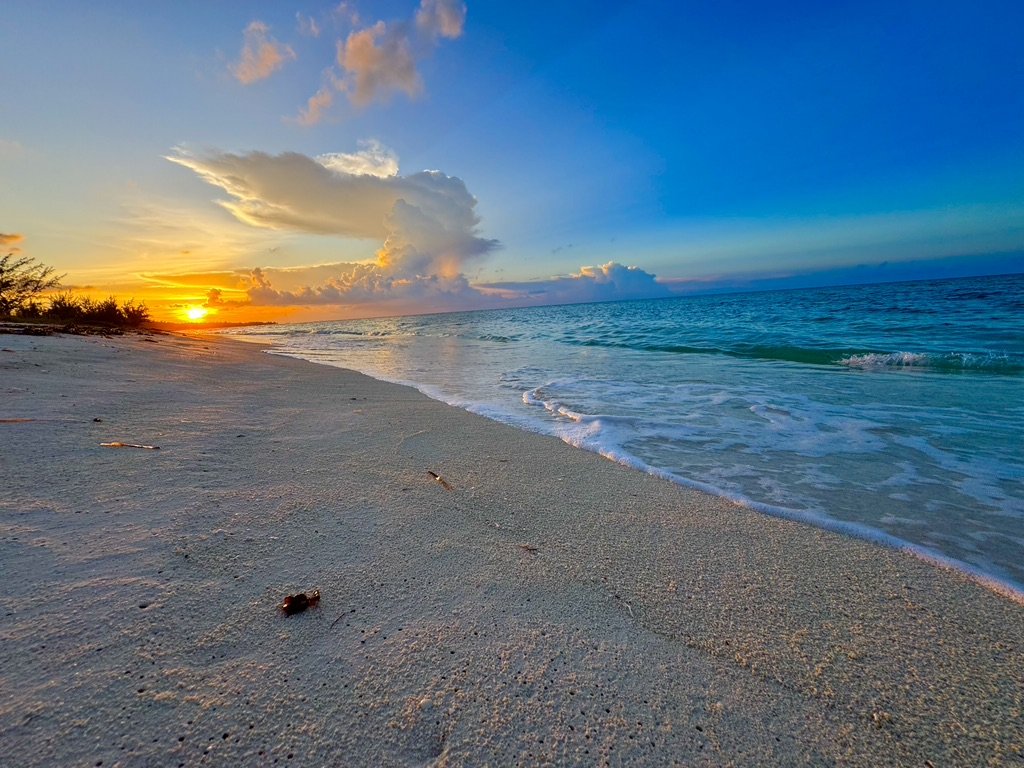 Sunset over a sandy beach with scattered small objects, calm ocean waves, and a partly cloudy sky.