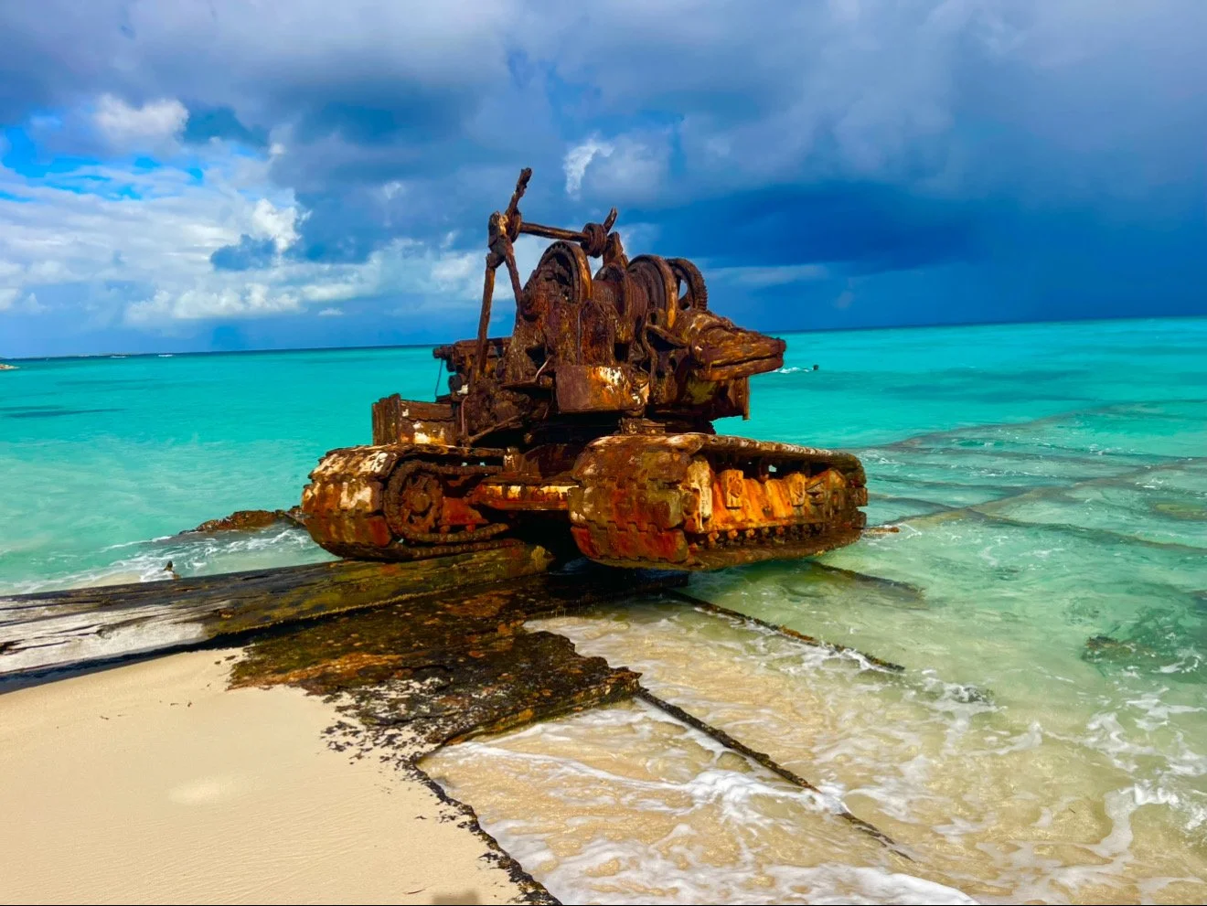 Rusty tank wreck on a sandy beach with clear turquoise water and a cloudy sky in the background.