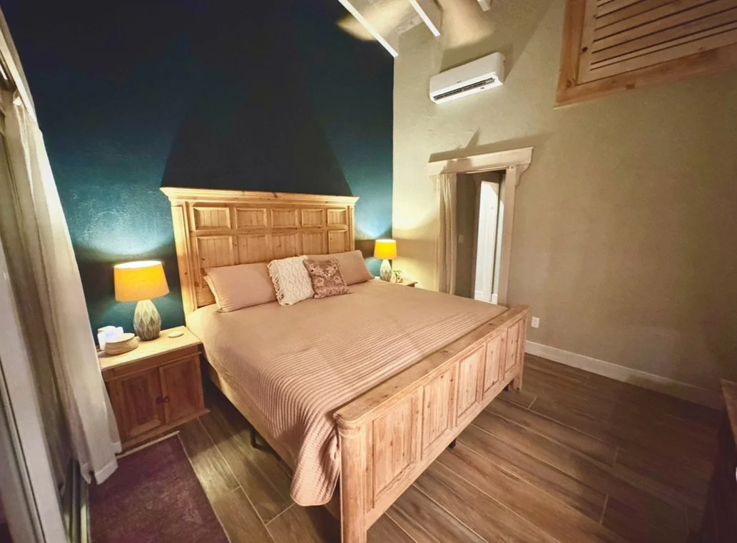 A cozy bedroom with a wooden bed frame, beige bedding, and pillows. Two nightstands with lamps are on each side of the bed. The room has dark blue and beige walls, wood flooring, and an air conditioning unit above a window.