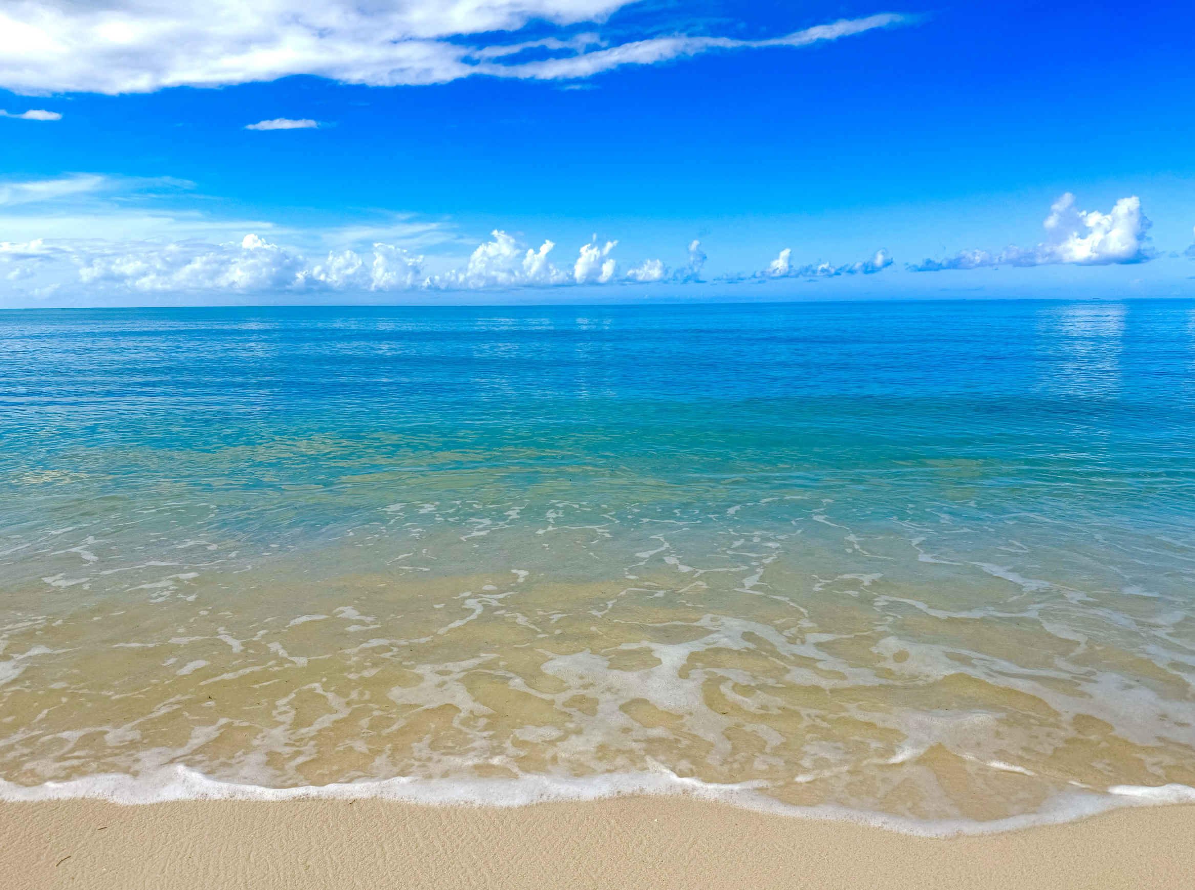 Beach scene with clear blue ocean water, sandy shore, and a sky with some clouds.