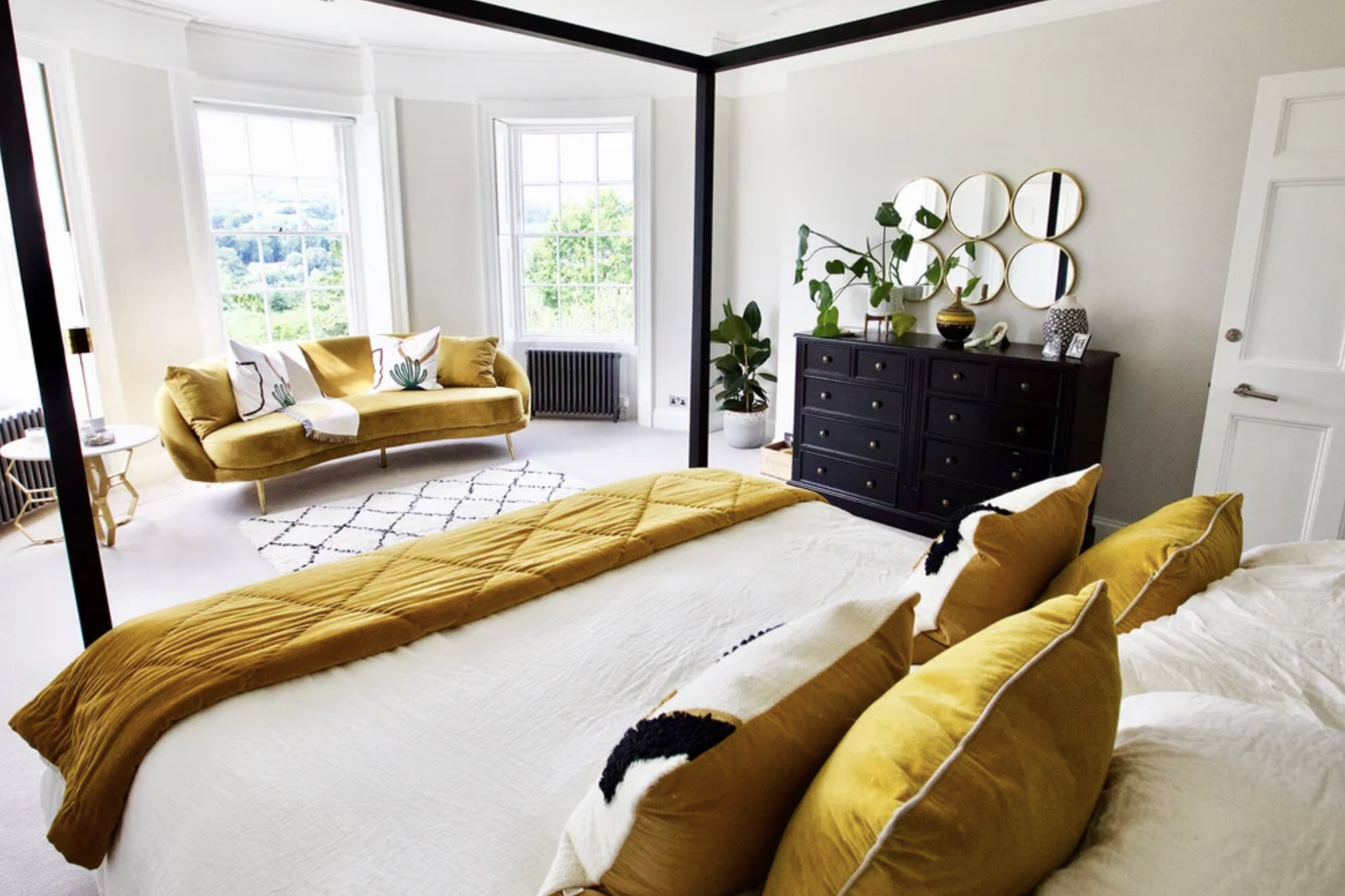 A bedroom with a large white bed, adorned with multiple gold pillows and a gold throw, facing a sitting area with a mustard-colored velvet sofa, white and green pillows, and a small side table. The room has three large windows with views of greenery outside, a black dresser with round mirrors above, and a potted plant in a white pot near the dresser. The room features neutral walls, white trim, and a beige carpet.