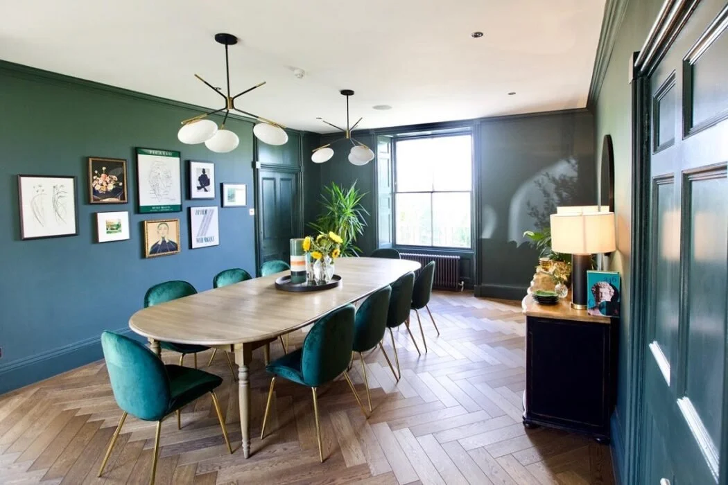 A stylish dining room with a long wooden table, surrounded by eight green velvet chairs with gold legs. The room features dark green walls decorated with framed artwork, a large window letting in natural light, and a black sideboard with a lamp, framed photo, and some decorative items.