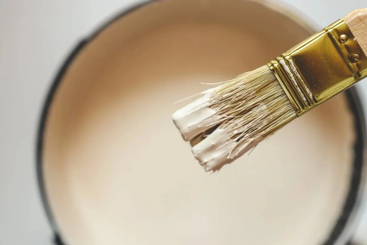 A paintbrush with white paint being dipped into a light-colored paint bucket.