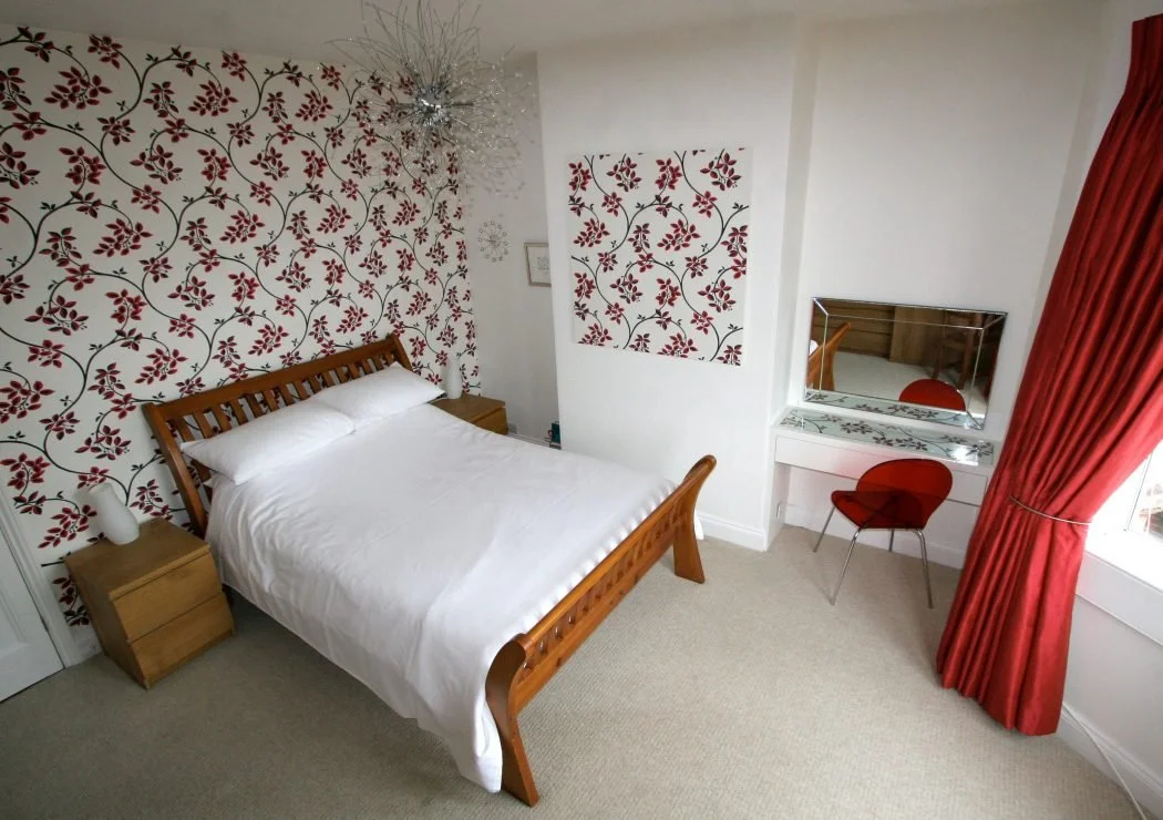 A bedroom with a bed, nightstands, patterned wallpaper, a mirror, a small desk, a red chair, and red curtains.