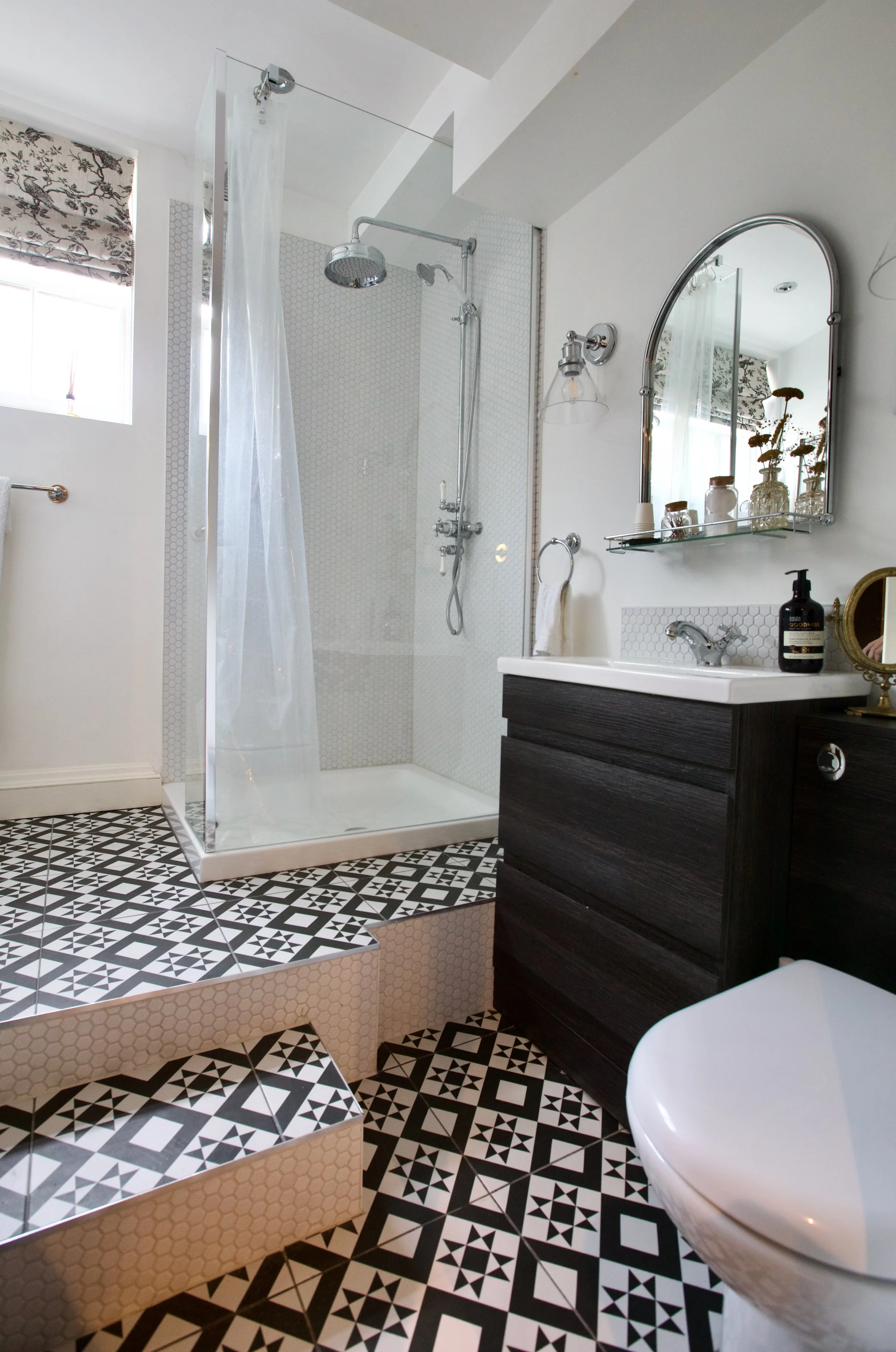 Modern bathroom with a patterned black and white tile floor, a walk-in shower with a glass door and white hexagon tiles, a black vanity with a white sink, an oval mirror, and decorative items on the shelf.