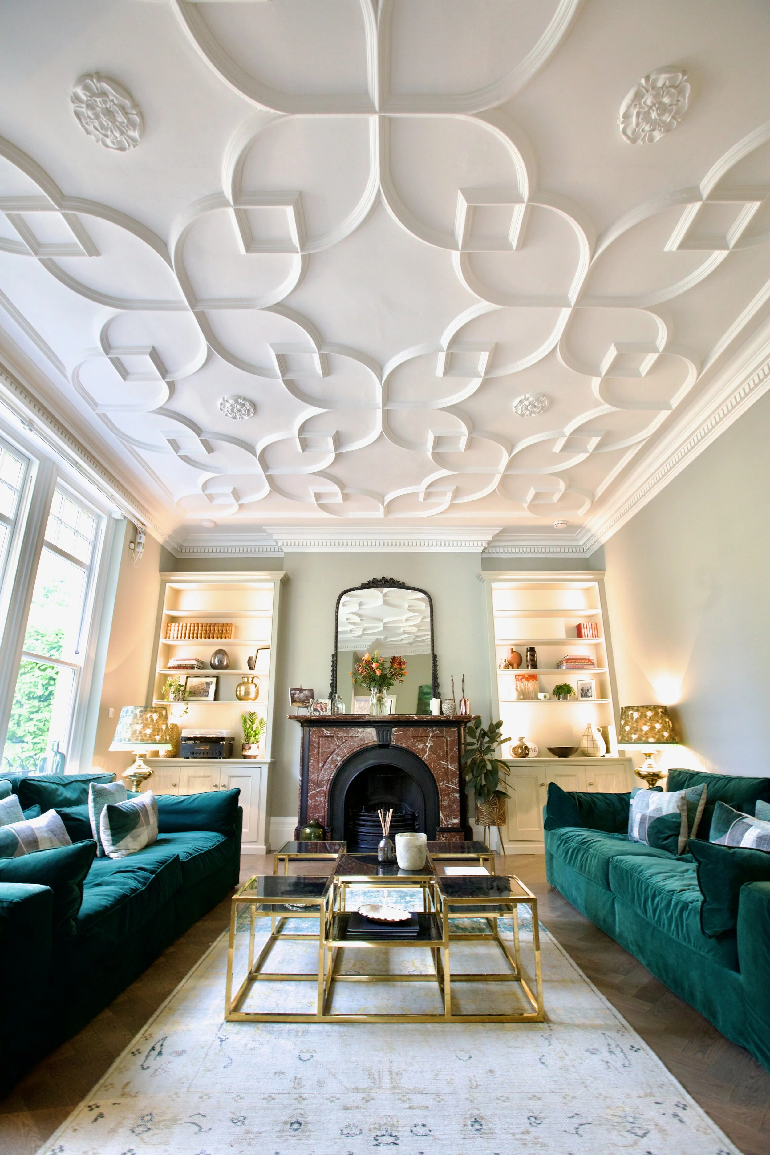 Living room with white decorative ceiling, green sofas, a fireplace with a mirror above, and built-in shelves with books and decor.