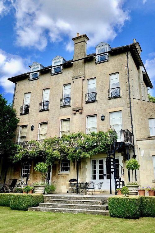 Four-story beige stone building with storm windows and small balconies, surrounded by greenery and outdoor furniture, under a partly cloudy sky.