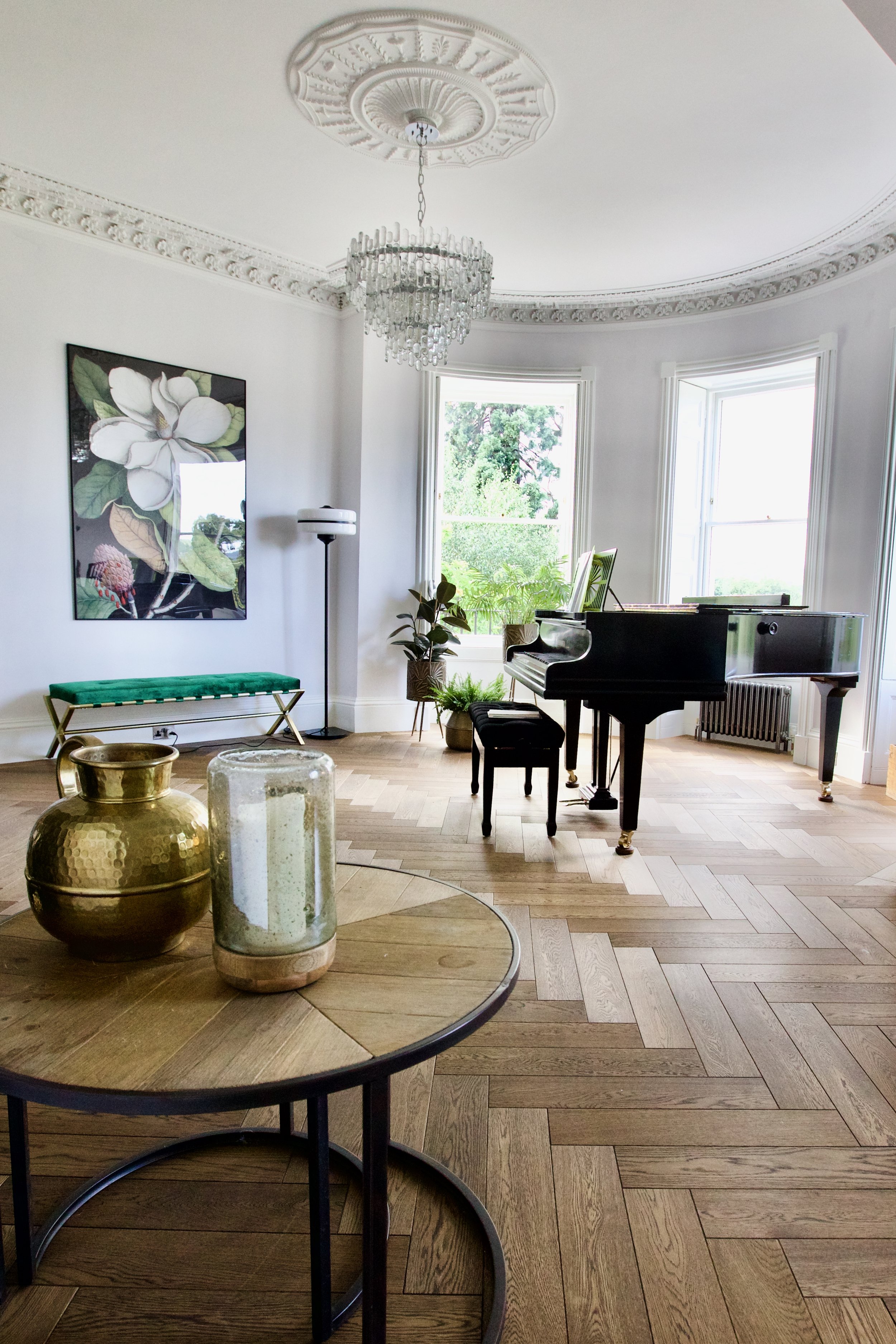 Living room with a grand piano, plants by the window, a chandelier, a floor lamp, a framed botanical print on the wall, and a wooden table with candles and a metallic vase.