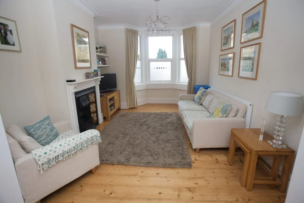 A bright living room with two cream-colored sofas, one with pillows and a throw blanket, a wooden side table with a lamp, a fireplace with a small television and shelves, a window with curtains, framed pictures on the walls, a chandelier, and a brown