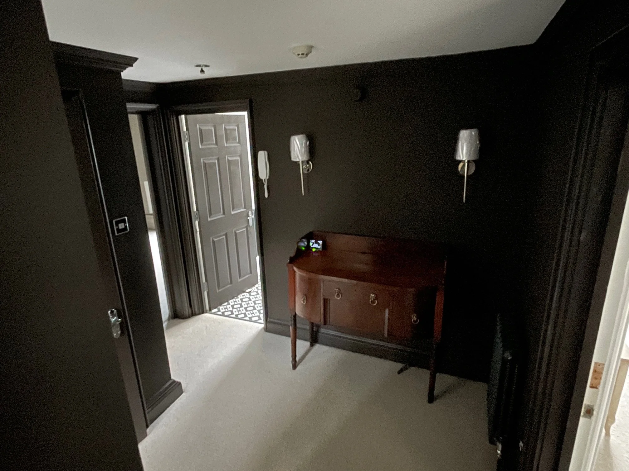 A dark interior room with light beige carpet, black walls, and black trim. There are two wall-mounted lamps with white shades, a small wooden side table with a phone and clock, and an open door leading outside, revealing a patterned black and white tile floor.