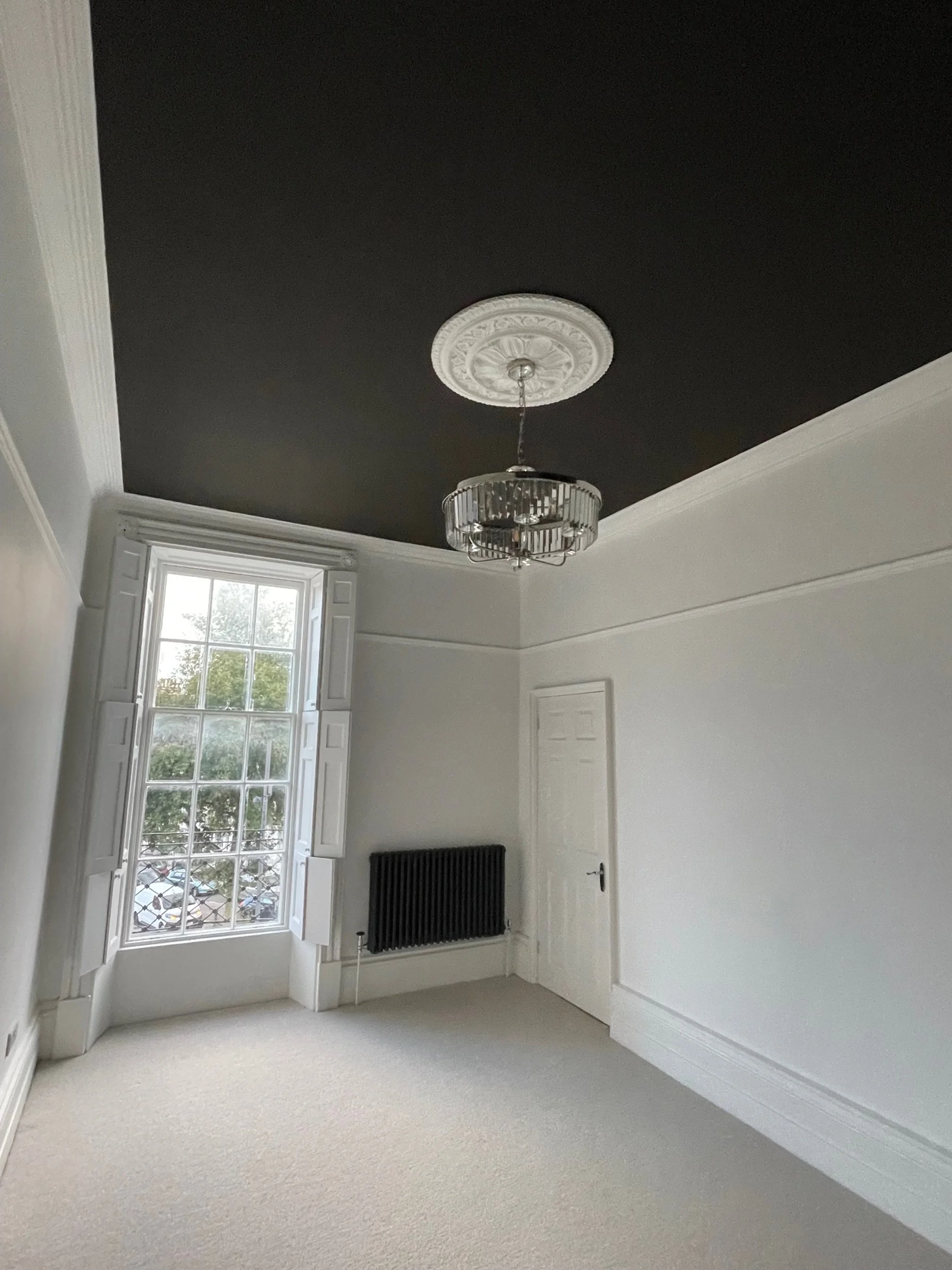 Empty room with white walls, a black ceiling, a decorative ceiling medallion, a hanging chandelier, a large window with shutters, and a radiator.