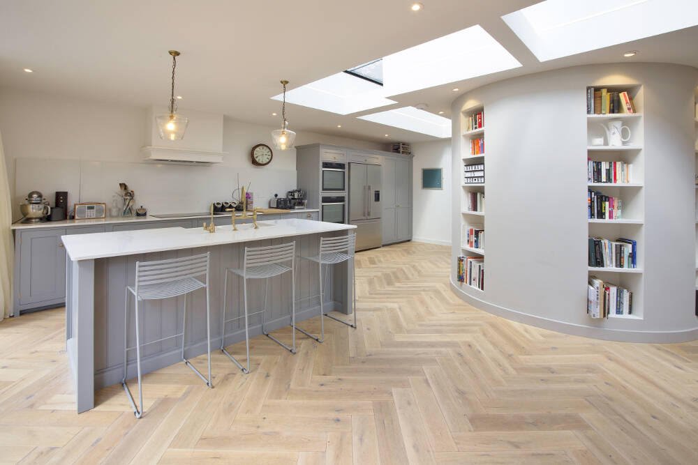 Modern kitchen with a white island, three white bar stools, built-in bookshelf with books, and skylights.