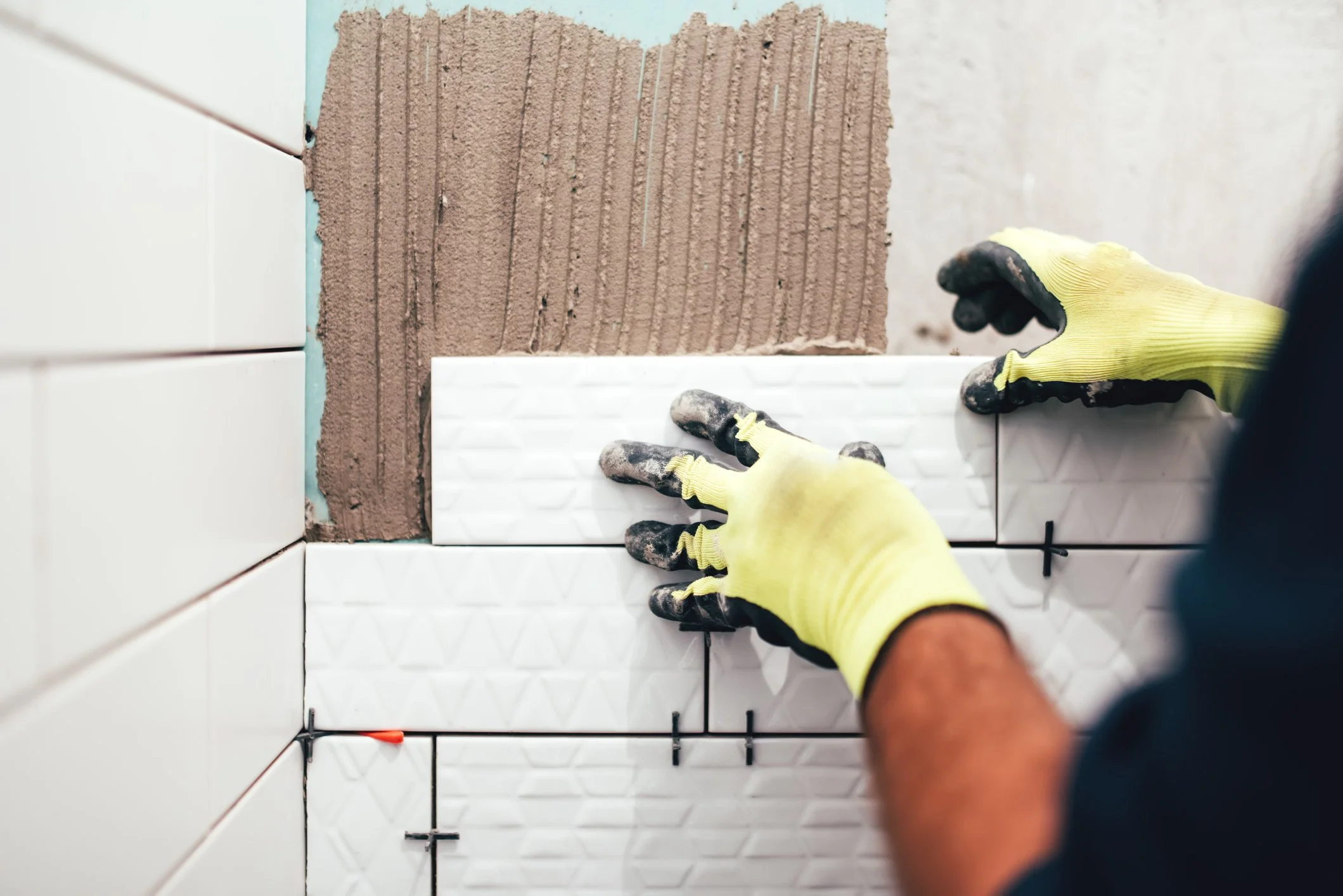 A person installing white textured ceramic tiles on a wall, with a section of brown adhesive and a blue wall behind.