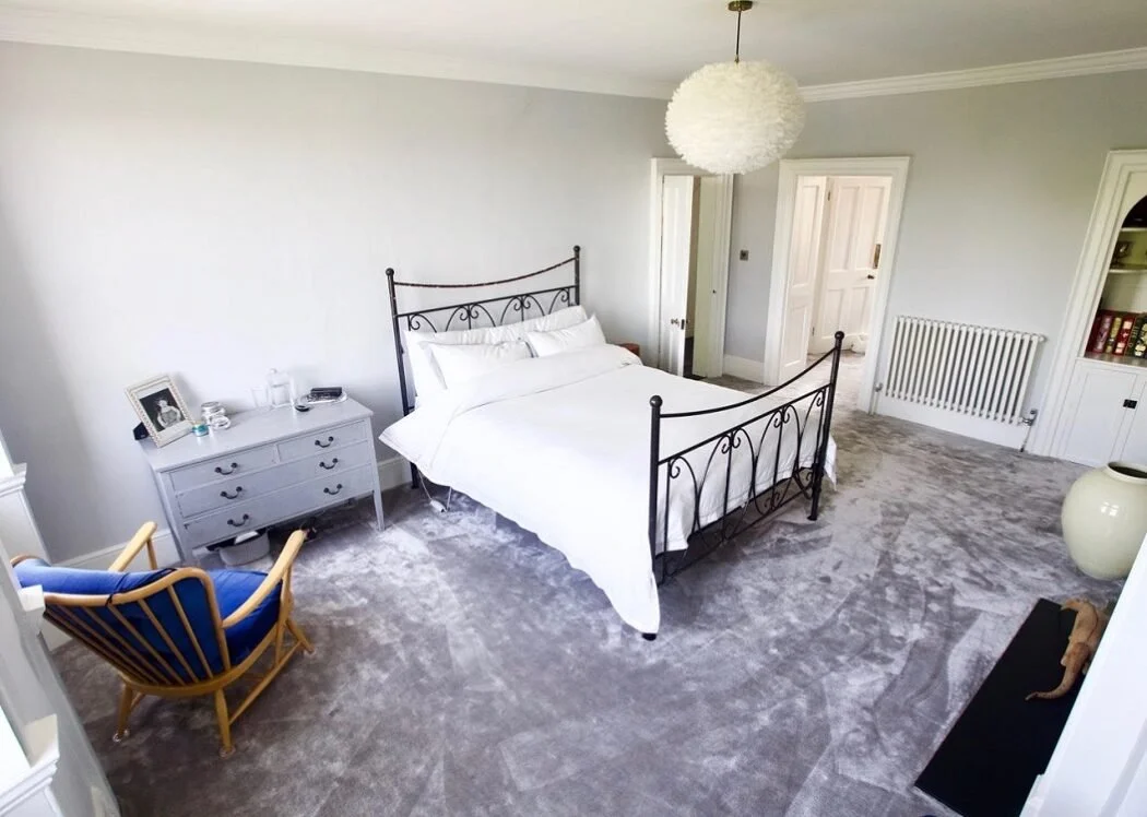 A bedroom with a bed, white bedding, and a dark metal frame, a blue and wooden rocking chair, a white side table with picture frame and glass, a large white vase, a black clothing rack, white walls, carpeted floors, a white lamp hanging from ceiling,