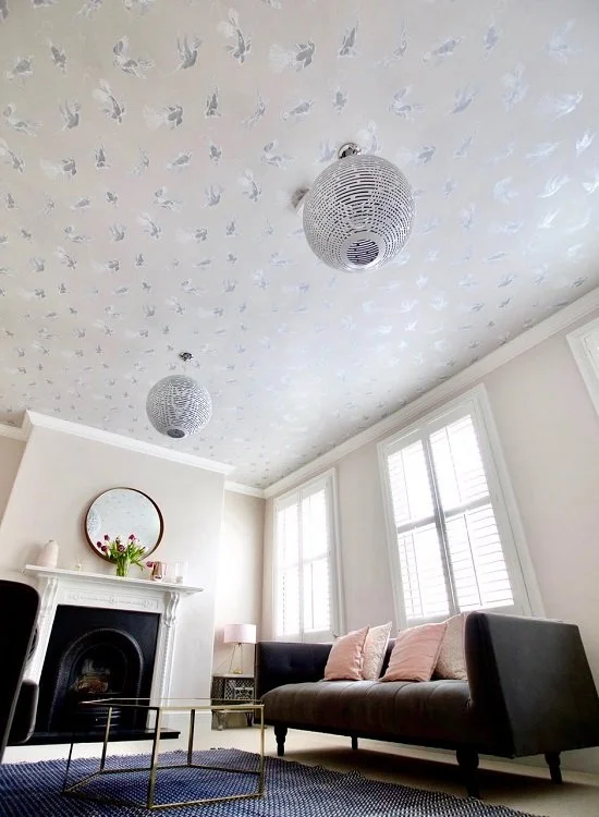 Living room with white walls, large windows with blinds, a dark gray sofa with pink pillows, white mantel with a mirror and vase of flowers, black fireplace, side table with lamp, and two hanging light fixtures in the ceiling with white dove patterns