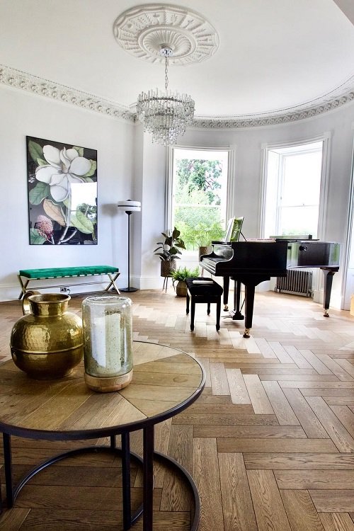 Room with a grand piano, chandelier, large windows, plants, portrait on the wall, and decorative vases on a table.