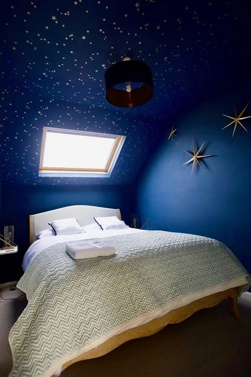 Bedroom with blue walls and ceiling decorated with white stars and gold star-shaped wall art, window on sloped ceiling, bed with white pillows and a zigzag patterned duvet, towel on bed, nightstand with lamp.