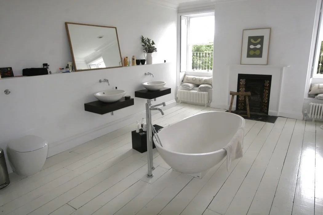 Bright bathroom with white wooden floors, a freestanding bathtub with a towel hanging over the edge, two floating sinks with a mirror above, a fireplace, and a window with a cushioned nook outside.