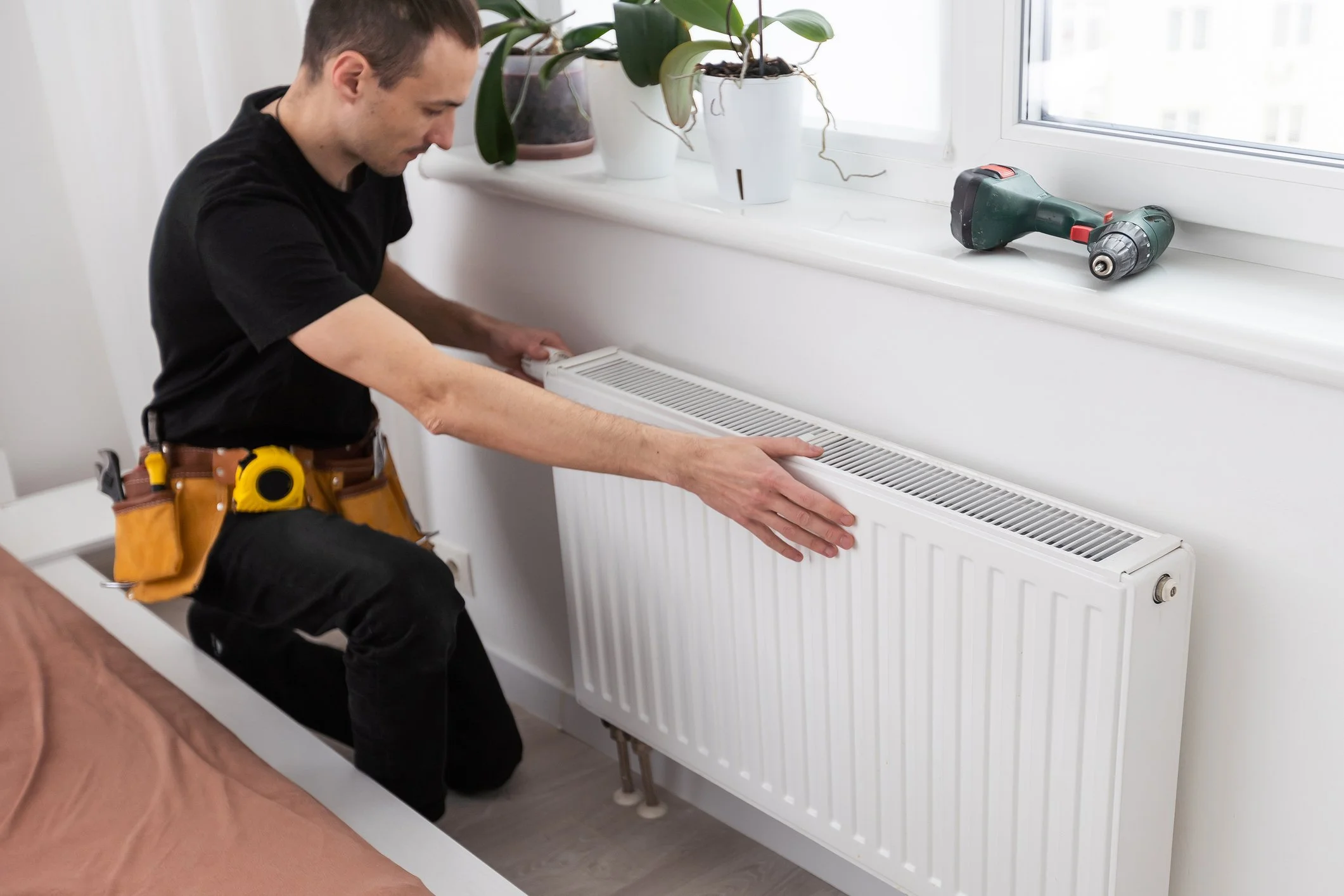 A man kneeling on a bed installing or inspecting a white radiator under a window, with tools and potted plants on the window sill.