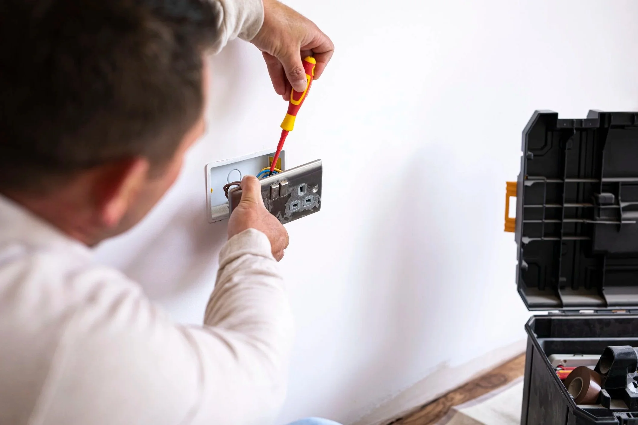 A person installing or repairing an electrical outlet using a screwdriver, with a toolbox nearby.