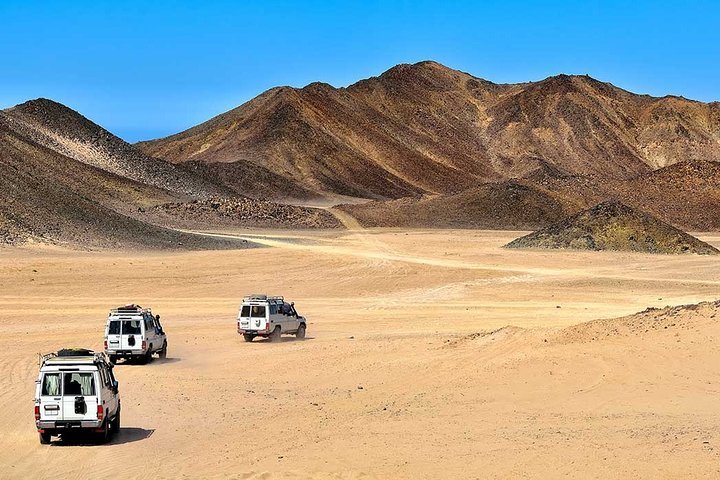 Three white off-road vehicles driving across a desert landscape with mountain ranges in the background under a clear blue sky.