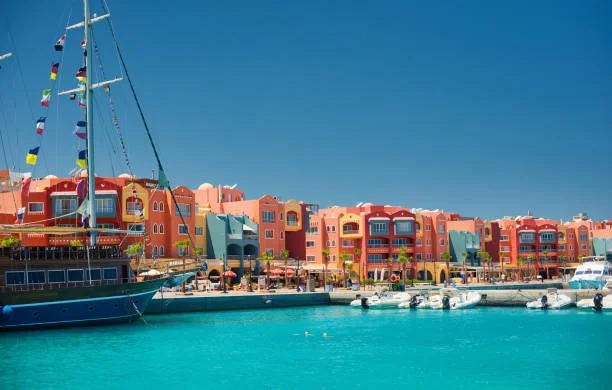 Colorful waterfront buildings with boats docked along the water, clear blue sky, and a large sailboat on the left.