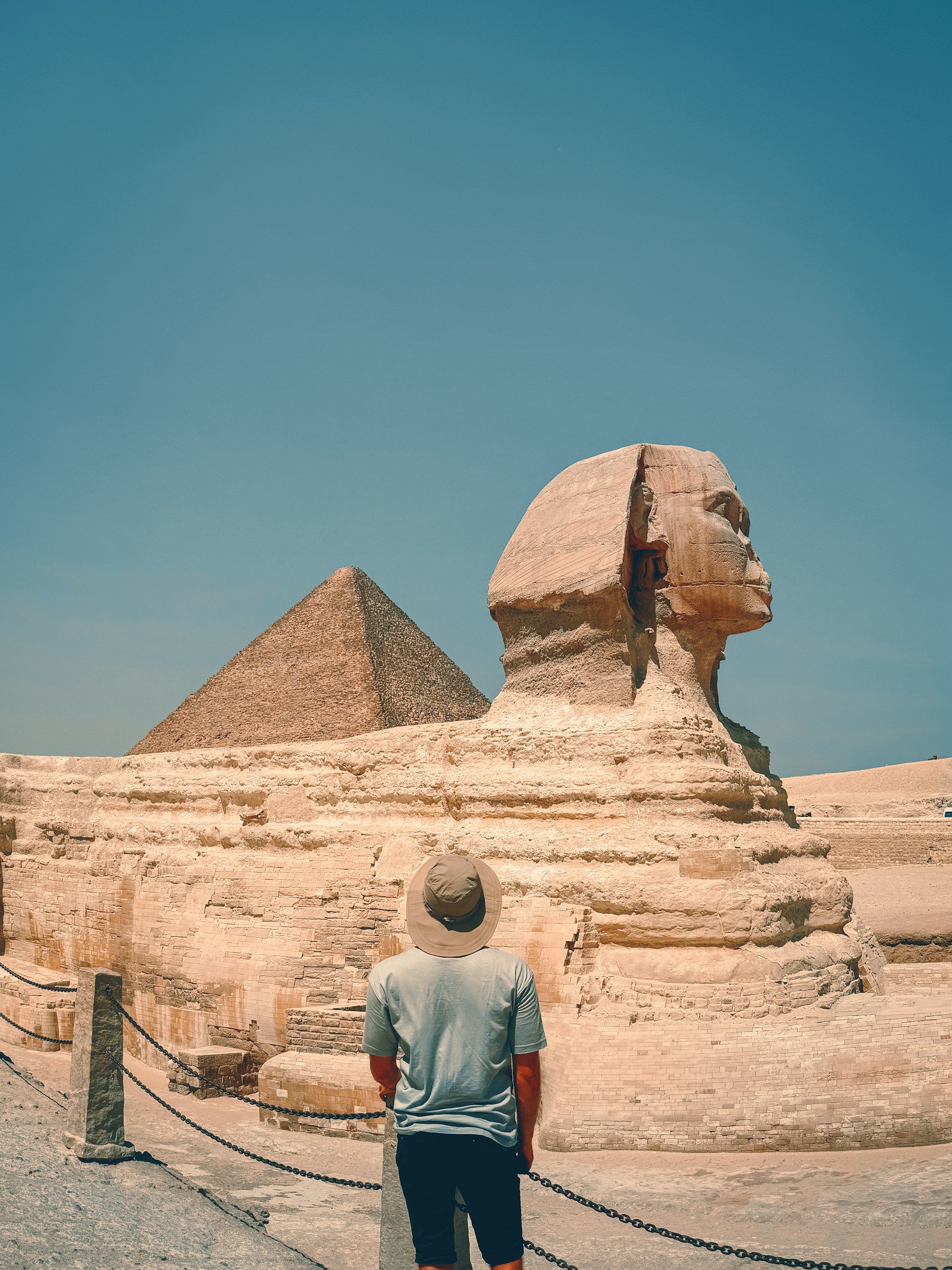 A person wearing a hat and t-shirt stands in front of the Great Sphinx of Giza with a pyramid in the background under a clear blue sky.