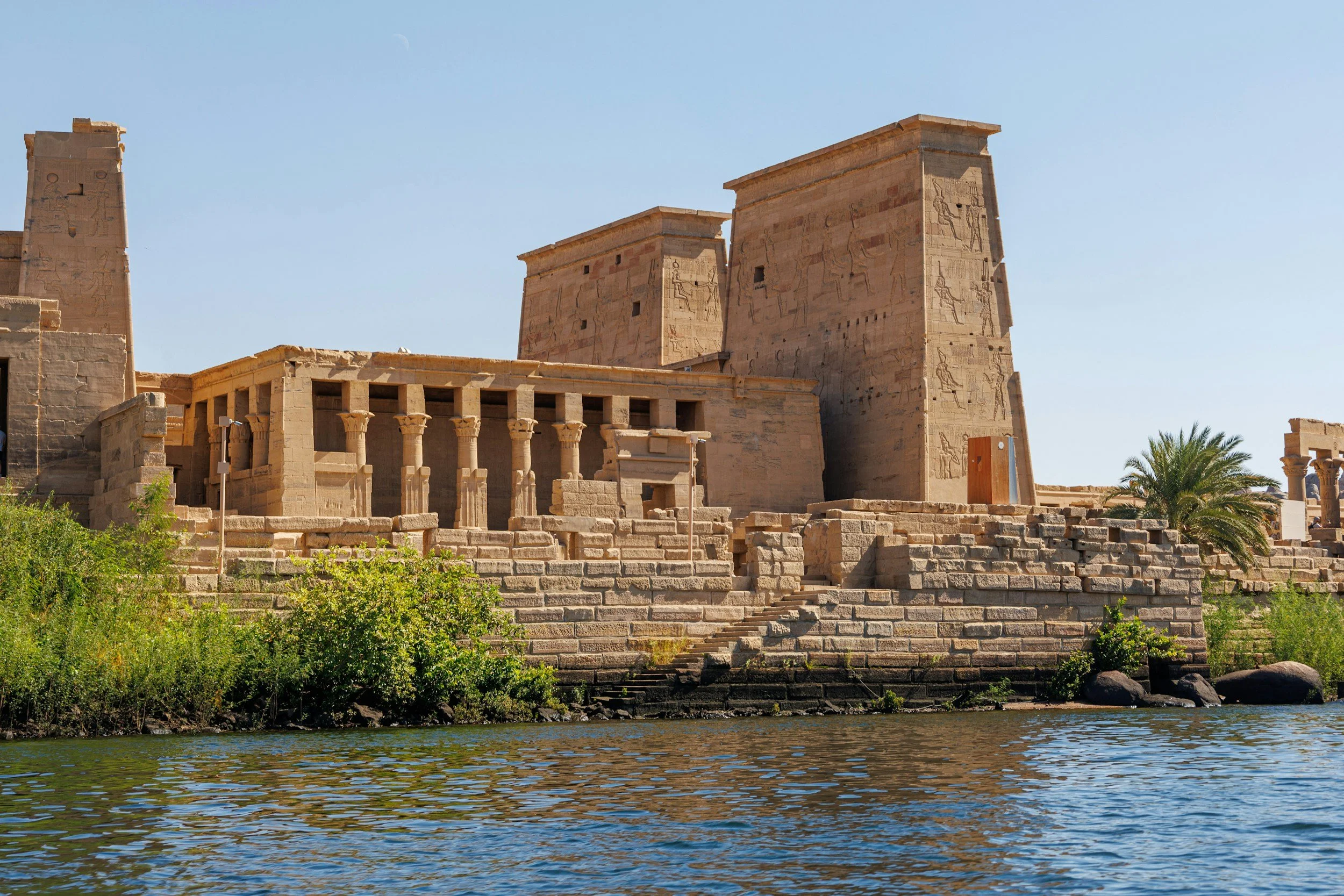 Ancient Egyptian temple ruins with large stone towers, columns, stairs, greenery, and water in the foreground under a clear blue sky.