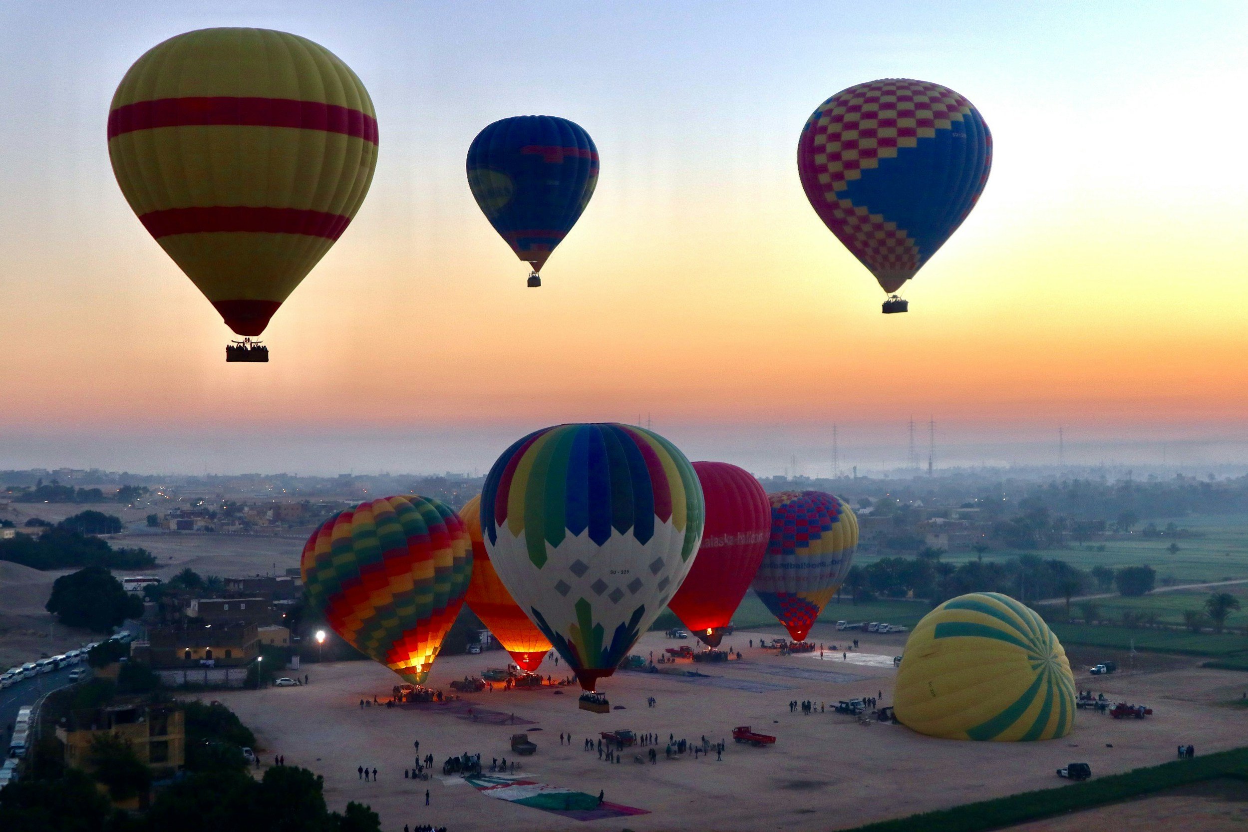 Multiple colorful hot air balloons flying at sunrise over a flat landscape, with some balloons on the ground preparing for flight.