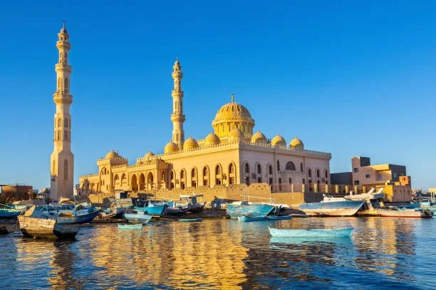 A mosque with two tall minarets, a large dome, and several smaller domes, situated by a harbor with boats, under a clear blue sky.