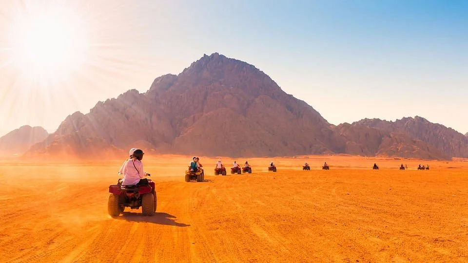A group of people riding all-terrain vehicles across a desert landscape with mountains in the background, under a bright sun.