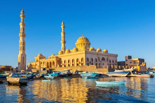 A mosque with multiple domes and tall minarets, situated by the water with numerous boats docked nearby under a clear blue sky.