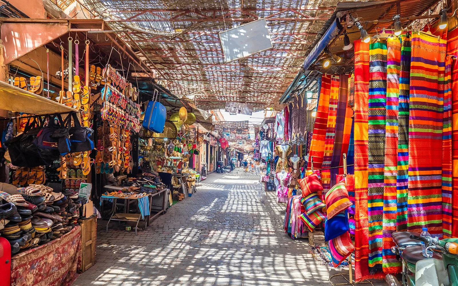 Colorful market street with stalls selling textiles, clothing, and accessories, with sunlight casting shadows through a woven roof.