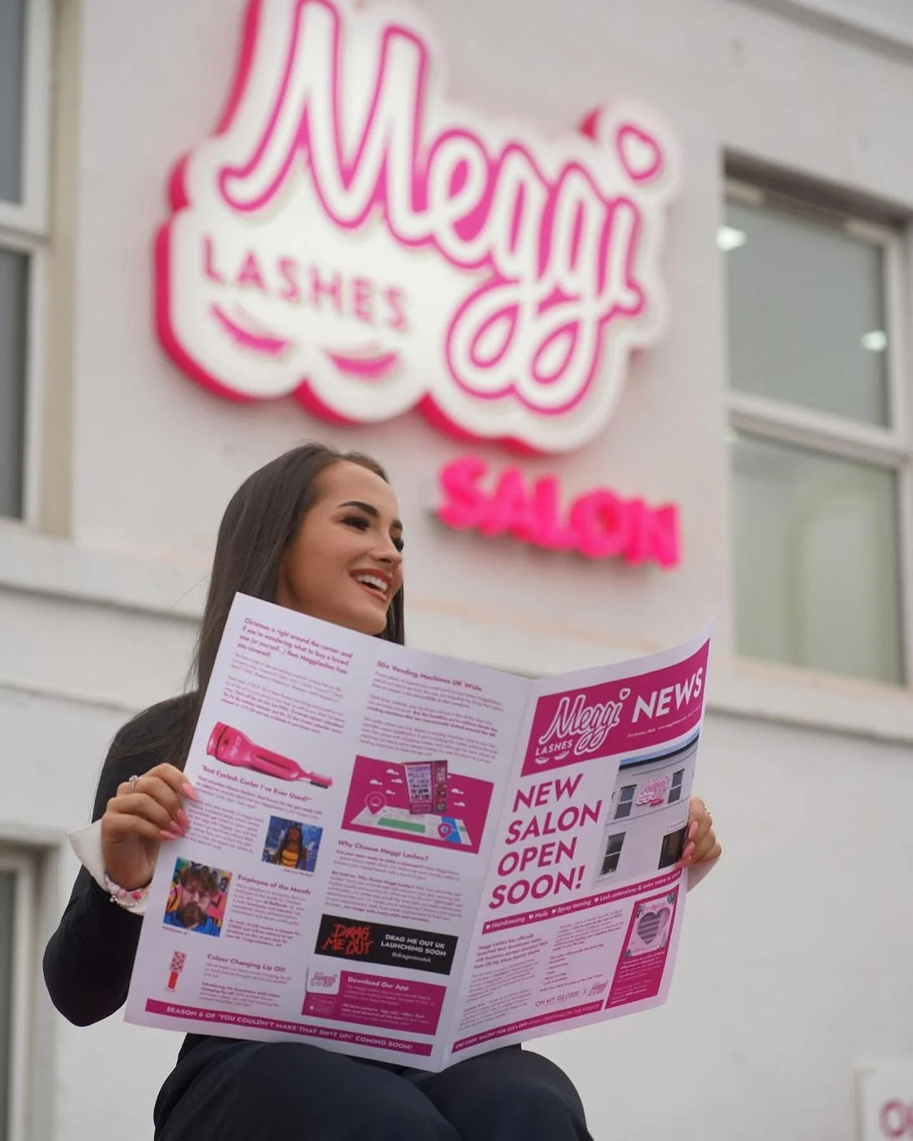 A woman with long dark hair smiling and holding a pink and white flyer outside a new salon with a sign that says 'Mega Lashes Salon' in bright pink letters.