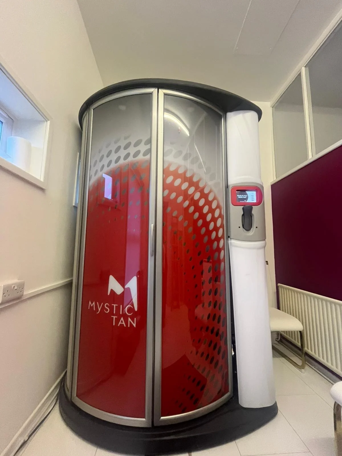 An outdoor sauna called Mystic Tan with a rounded glass door, red interior, and a white control panel on the side.