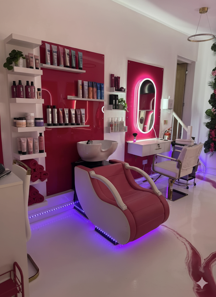 A modern beauty salon or spa treatment room with pink and white decor. Contains a reclining chair, a sink, shelves with hair and skincare products, a mirror with LED lighting, and a white chair. There are pink towels and purple LED lighting under the