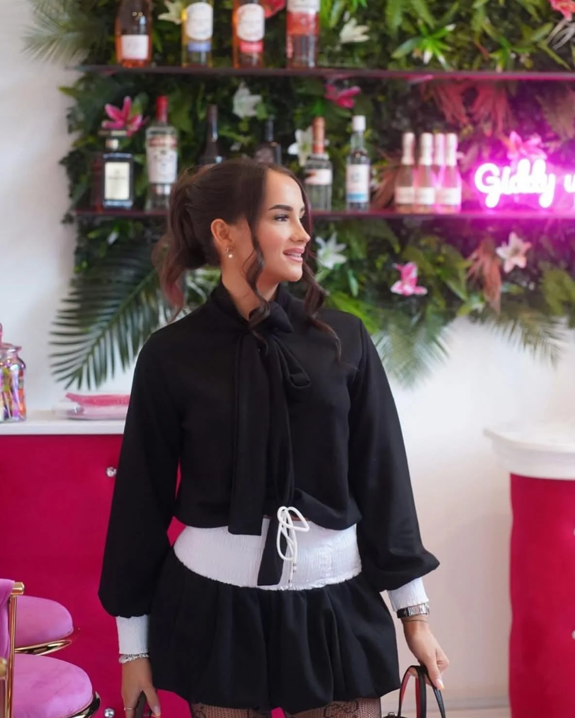A woman with dark wavy hair styled in loose curls wearing a black blouse with a bow tie at the collar, a white waistband, and a black skirt, standing indoors in front of a bar with bottles and a neon sign that reads 'Giddy up'. The background feature