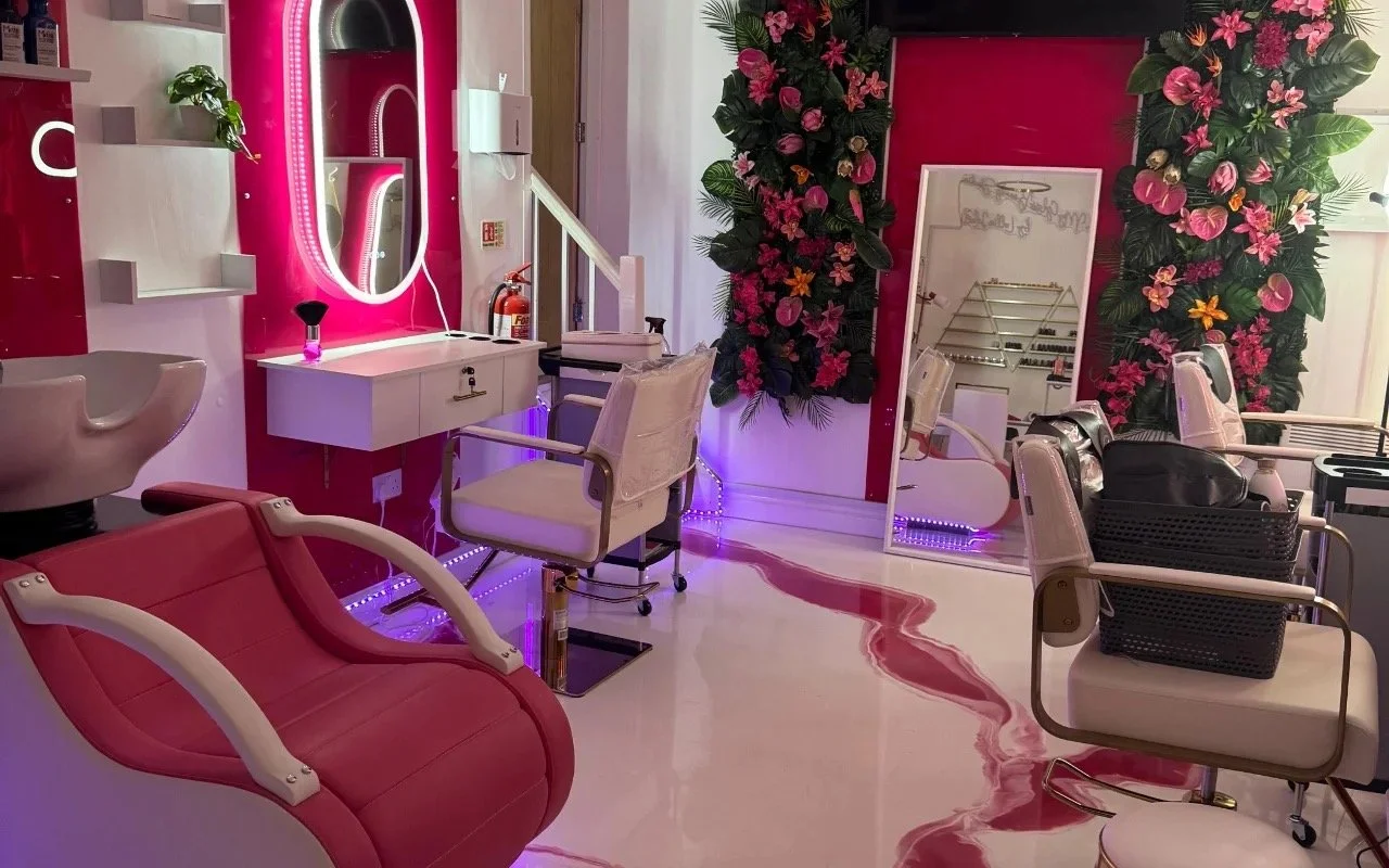 Inside a modern hair salon with pink and white decor, featuring styling chairs, a large mirror, decorative floral arrangements, and purple lighting accents.
