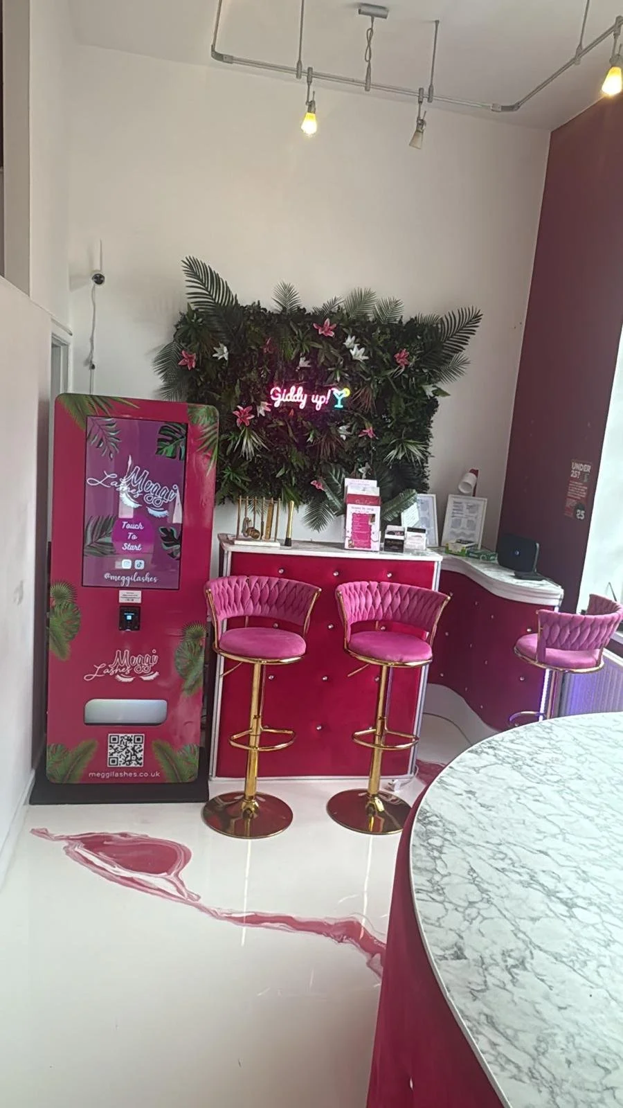 Indoor scene with pink bar stools, a pink and gold bar counter, a floral wall decoration with a neon sign that says 'Giddy up!', and a pink vending machine labeled 'Megglashs' with tropical leaf prints.