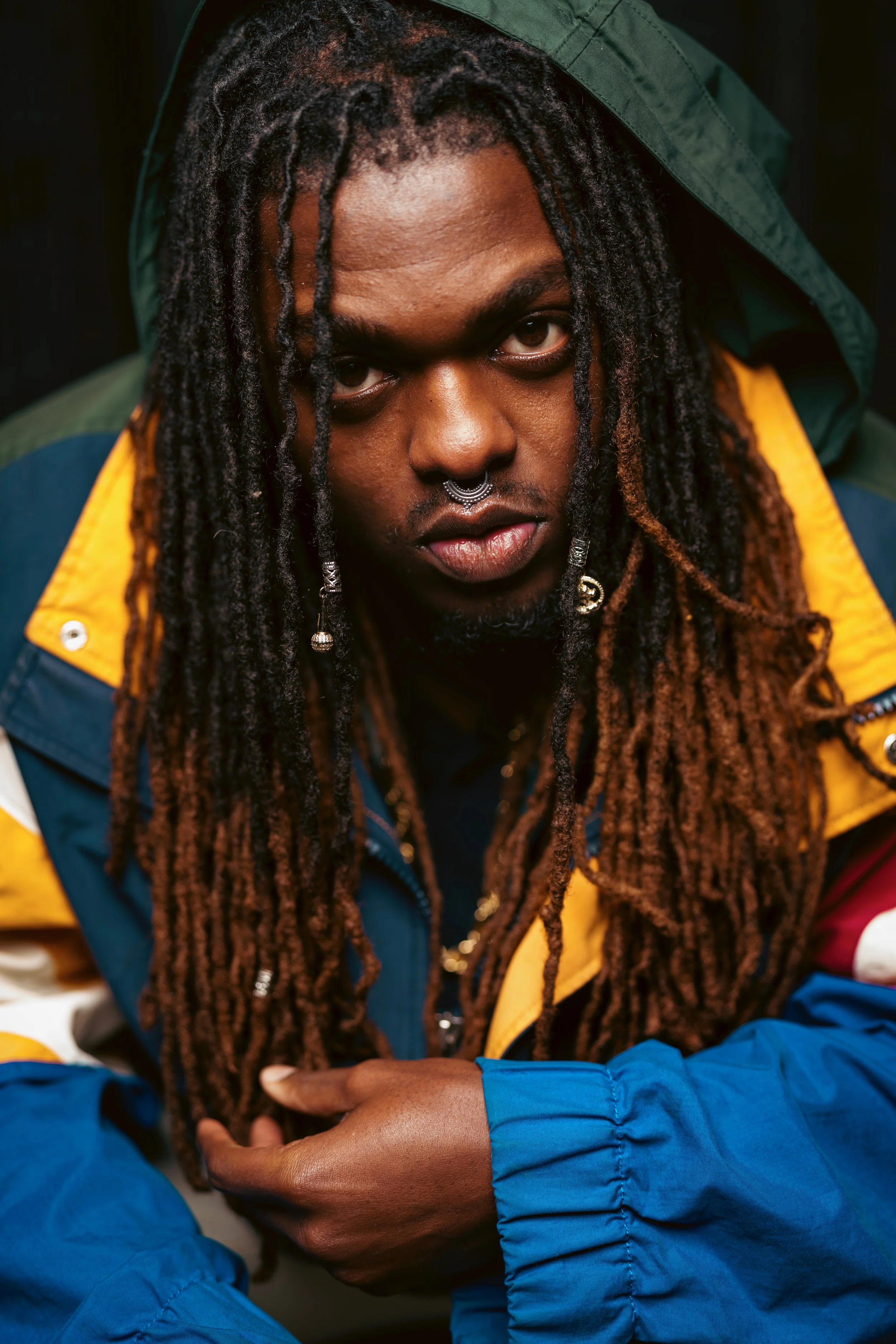 A person with long dreadlocks, a septum piercing, and earrings, wearing a colorful jacket with yellow, green, red, blue, and white sections, looking directly into the camera.