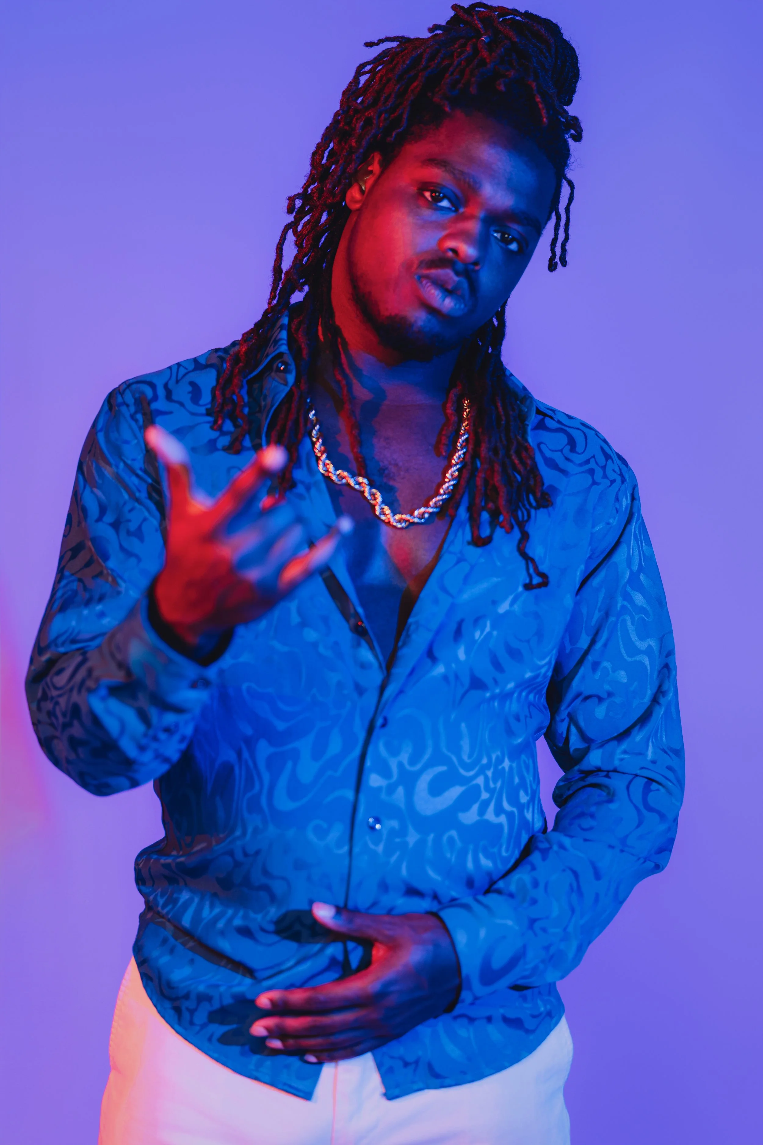 A man with long dreadlocks wearing a patterned blue shirt and white pants, posing with a hand gesture against a purple background with colorful lighting.