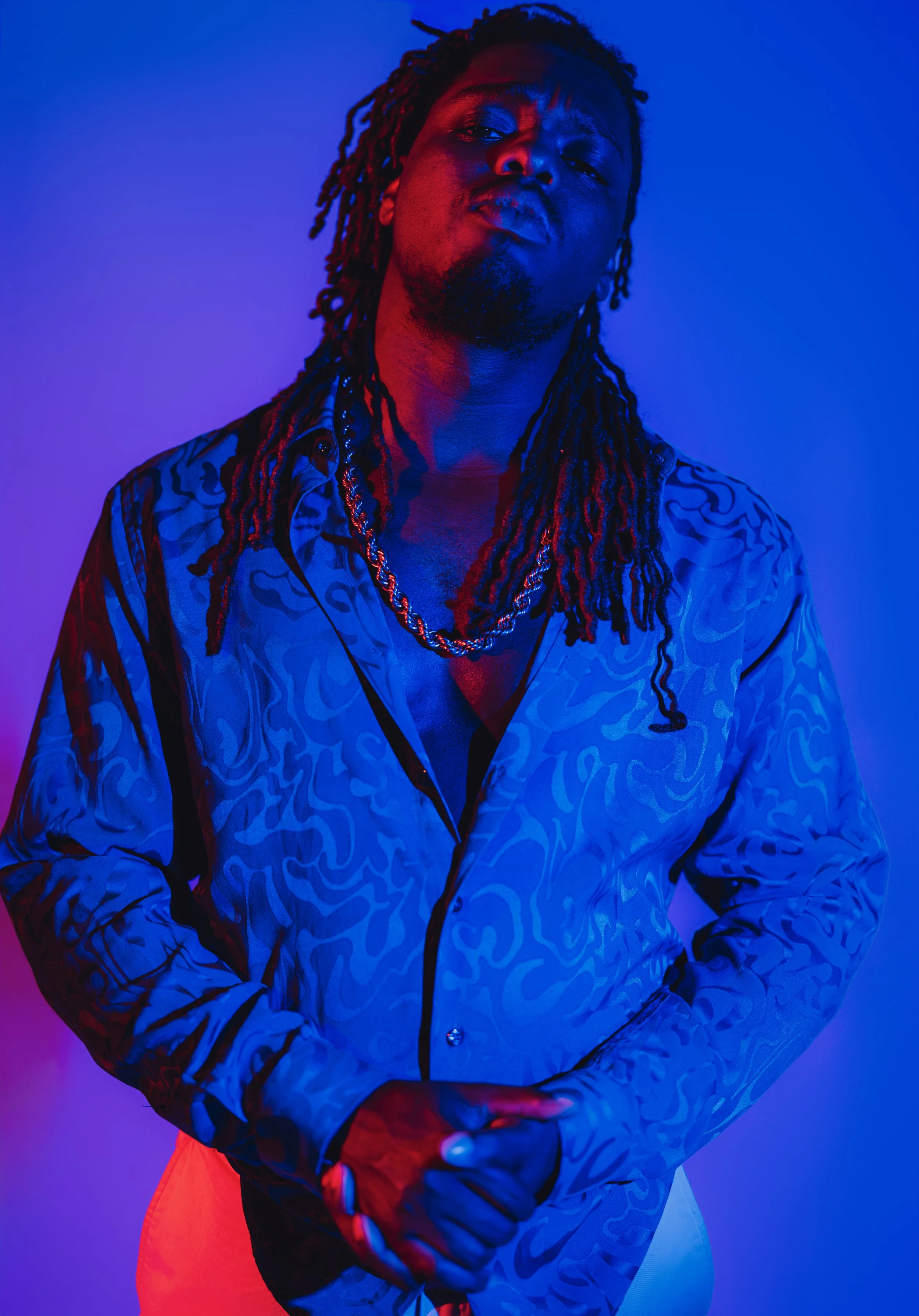 A man with dreadlocks and a chain necklace posing against a purple and blue background, wearing a shiny patterned shirt.