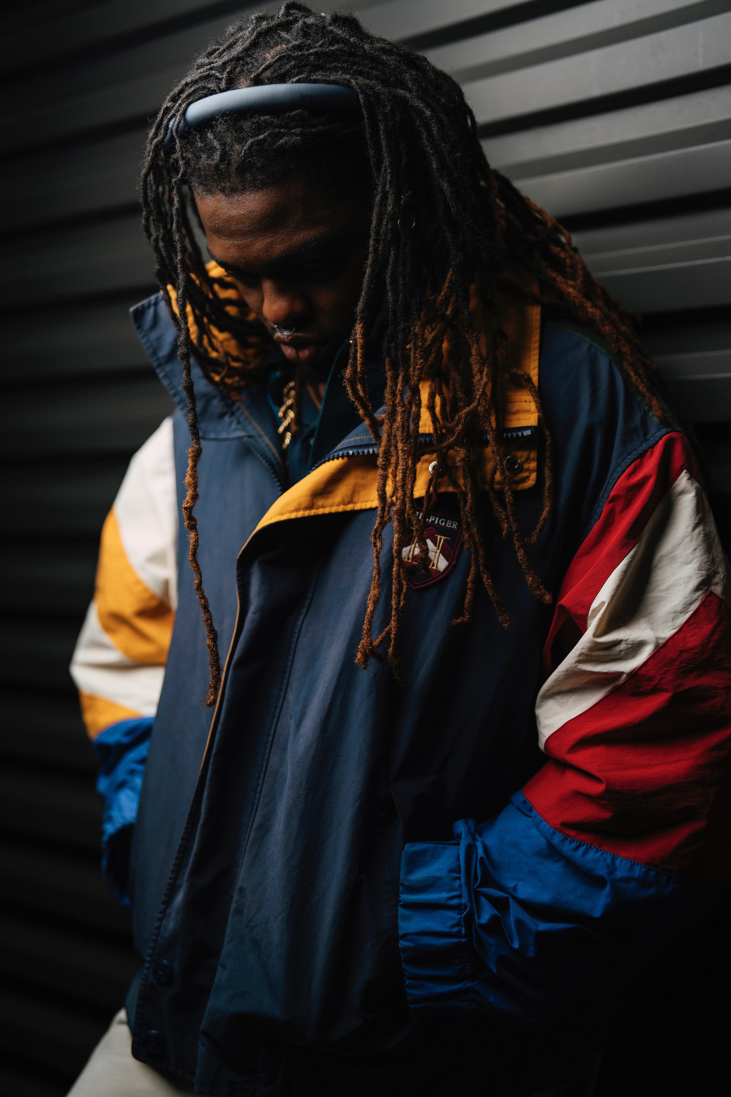 A man with dreadlocks wearing a colorful Tommy Hilfiger jacket, standing against a dark, corrugated metal wall, looking downward.