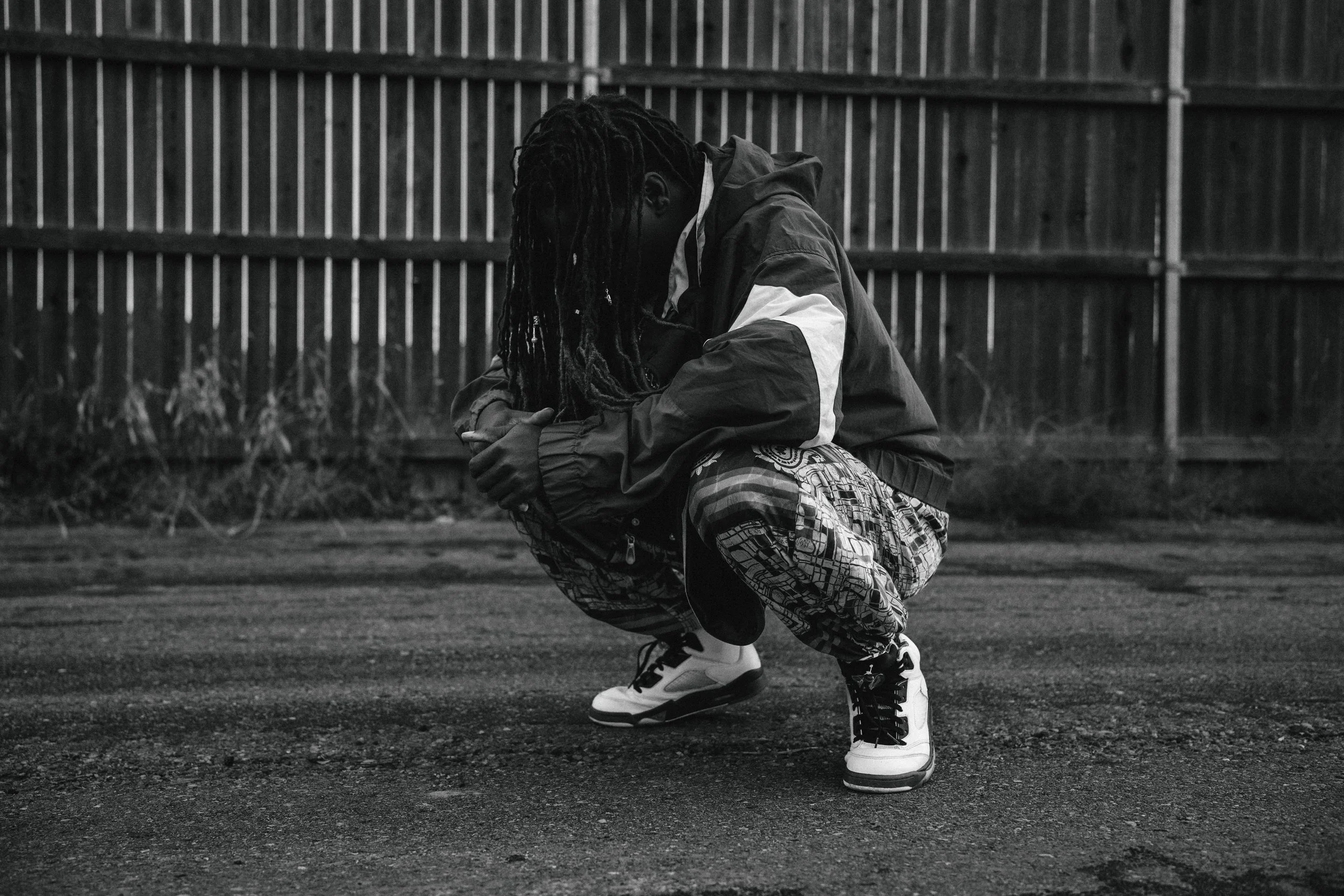 A person squatting outdoors near a wooden fence, dressed in a windbreaker jacket, patterned pants, and sneakers, with long dreadlocked hair.