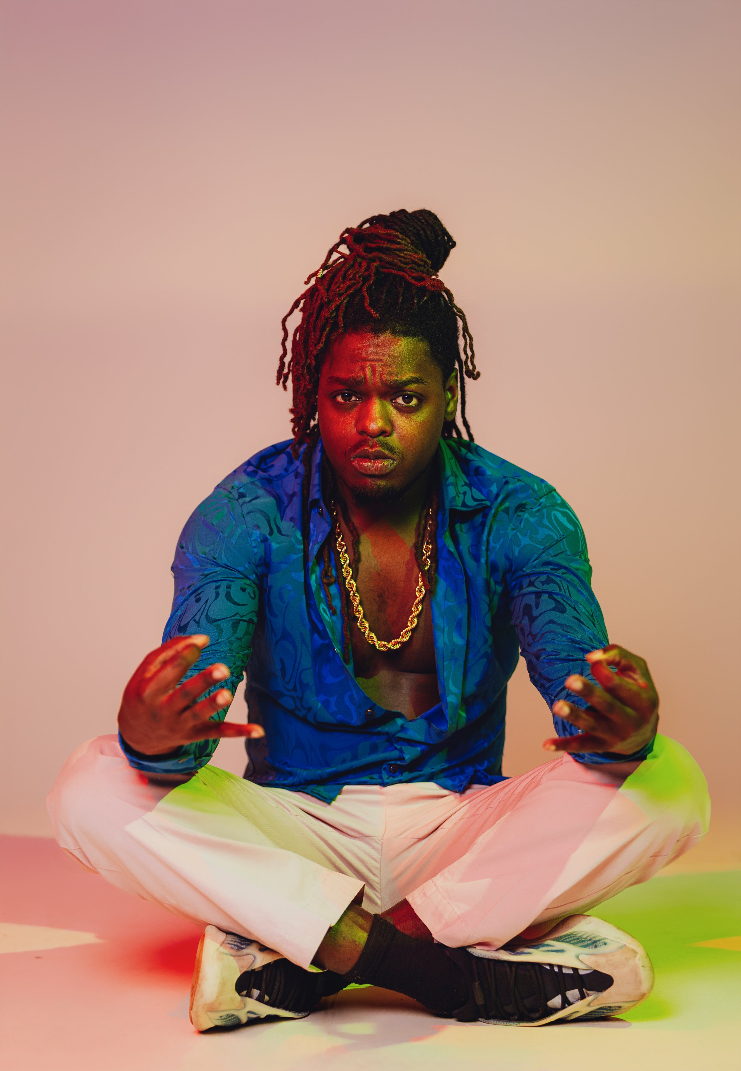A young man with dreadlocks tied up, sitting cross-legged on the floor, wearing a blue patterned shirt open to reveal a gold chain, white pants, and black sneakers. He has a serious expression against a plain background with colorful lighting effects.