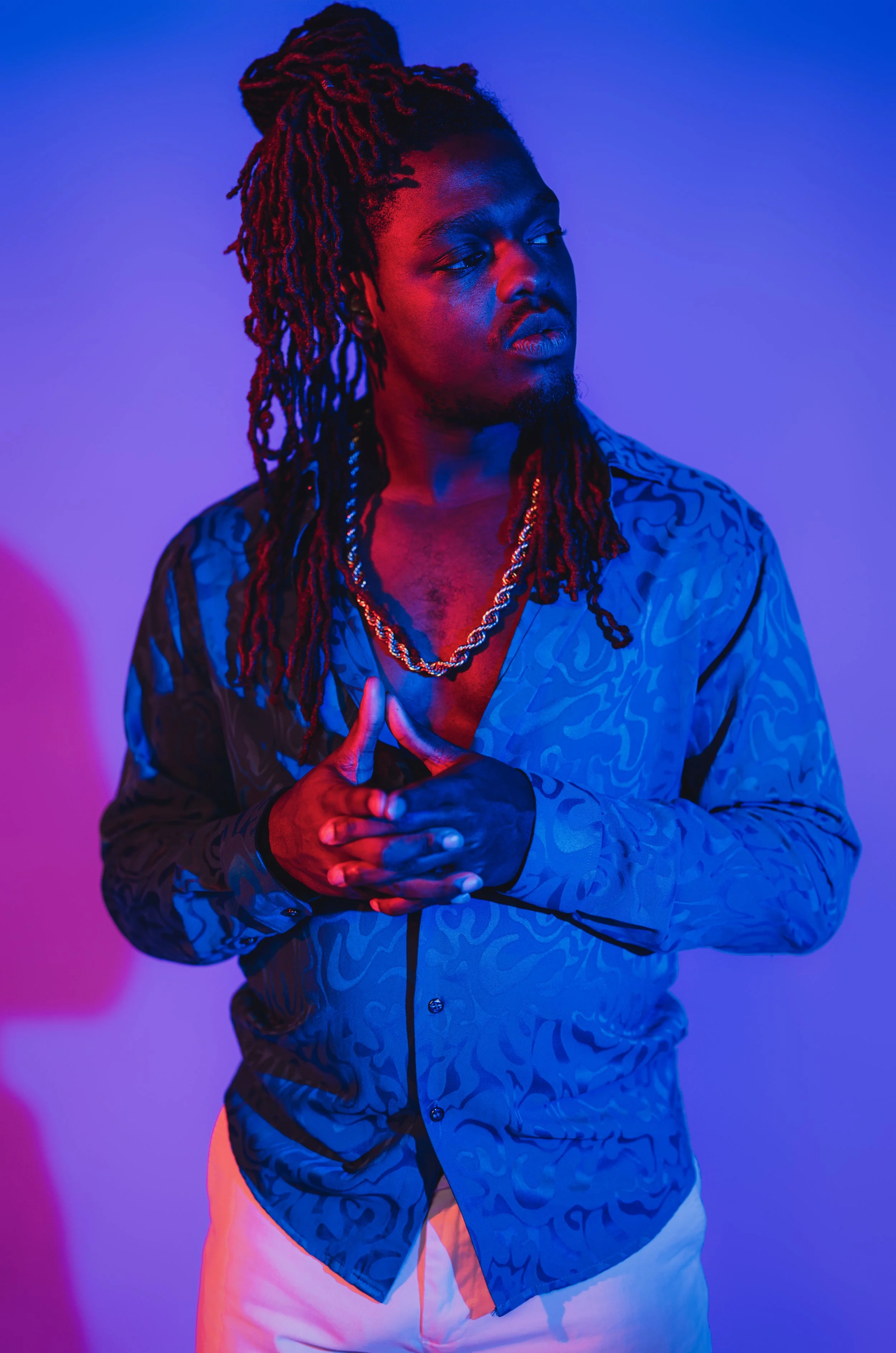A man with long dreadlocks and a goatee, wearing a patterned blue shirt, gold chains, and light-colored pants, poses with hands clasped in front of his chest against a background with purple and pink lighting.