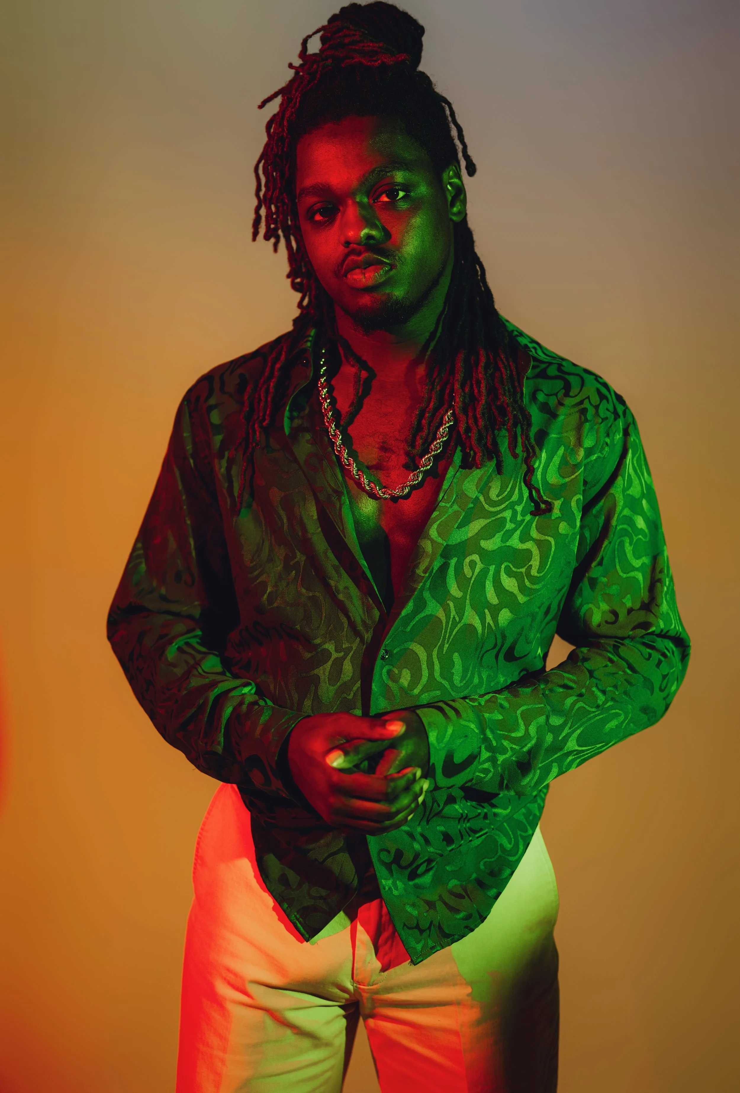 A young man with long dreadlocks wearing a patterned green shirt, light-colored pants, gold chains, and standing against a gradient background with red and green lighting.