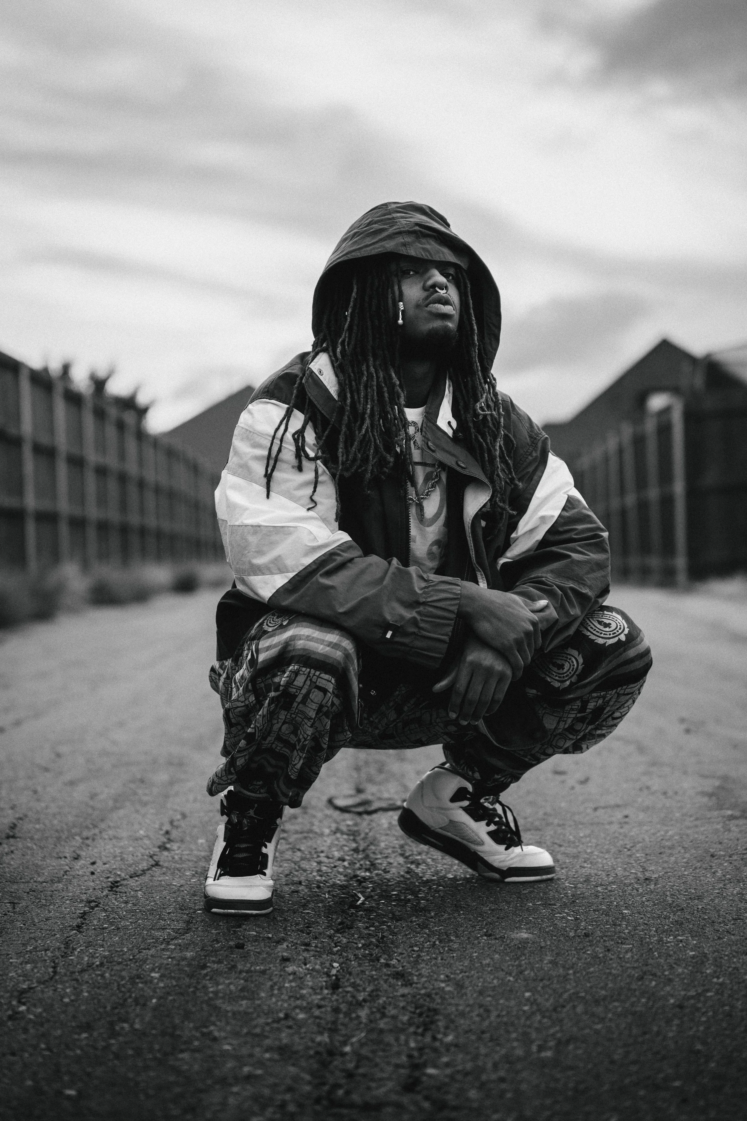 A man with dreadlocks squatting on a street, wearing a hoodie, a patterned jacket, patterned pants, and sneakers, with houses in the background and an overcast sky.